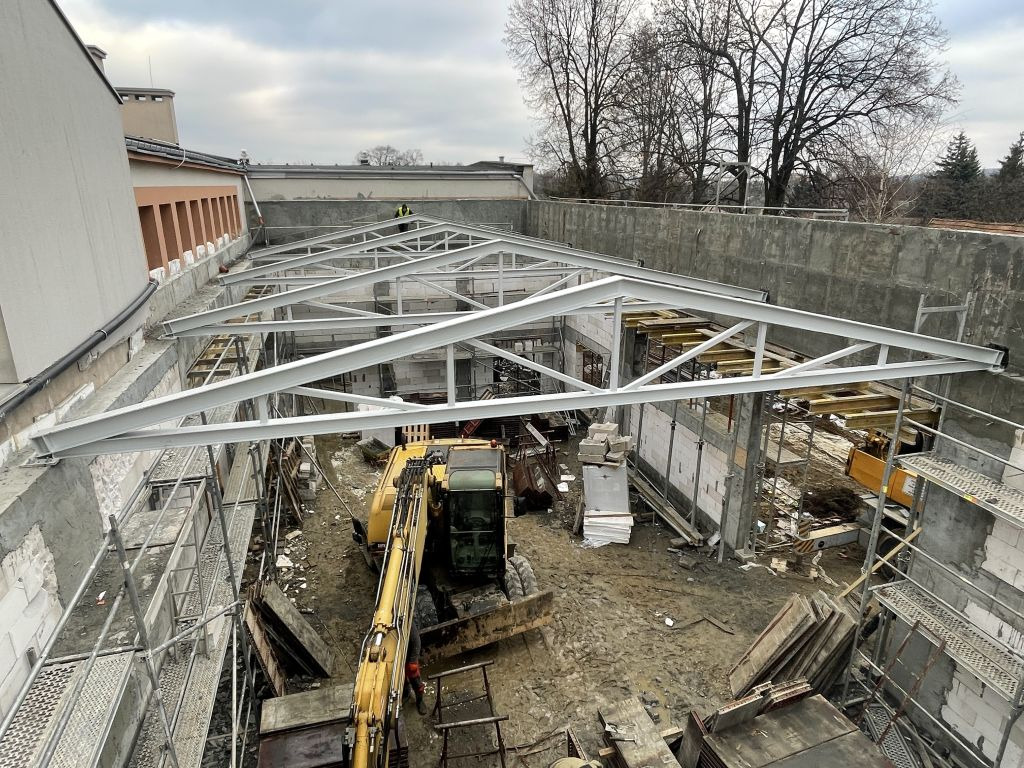 Budowa hali: stalowe konstrukcje z żółtą koparką na placu budowy, widok z góry na szare niebo.