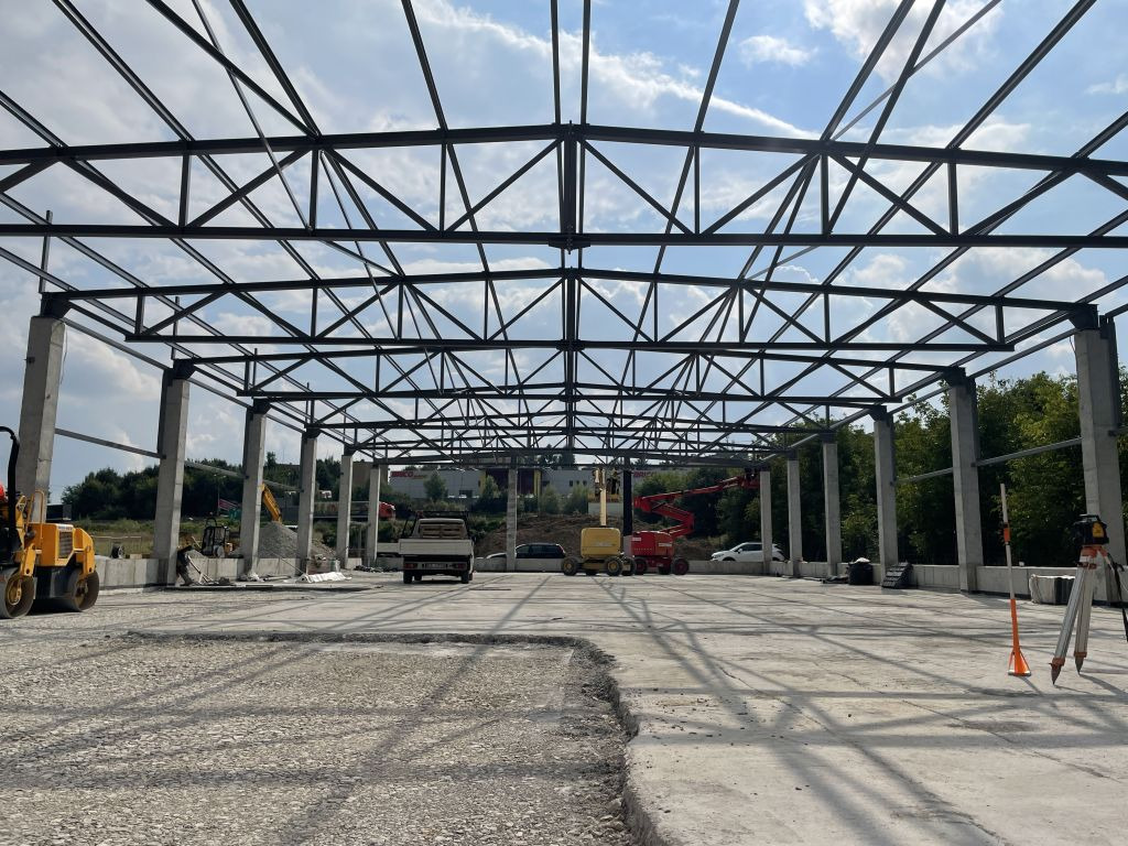 Konstrukcja stalowa hali w trakcie budowy z widocznymi słupami betonowymi, stalową kratownicą dachu i maszynami budowlanymi w tle.
