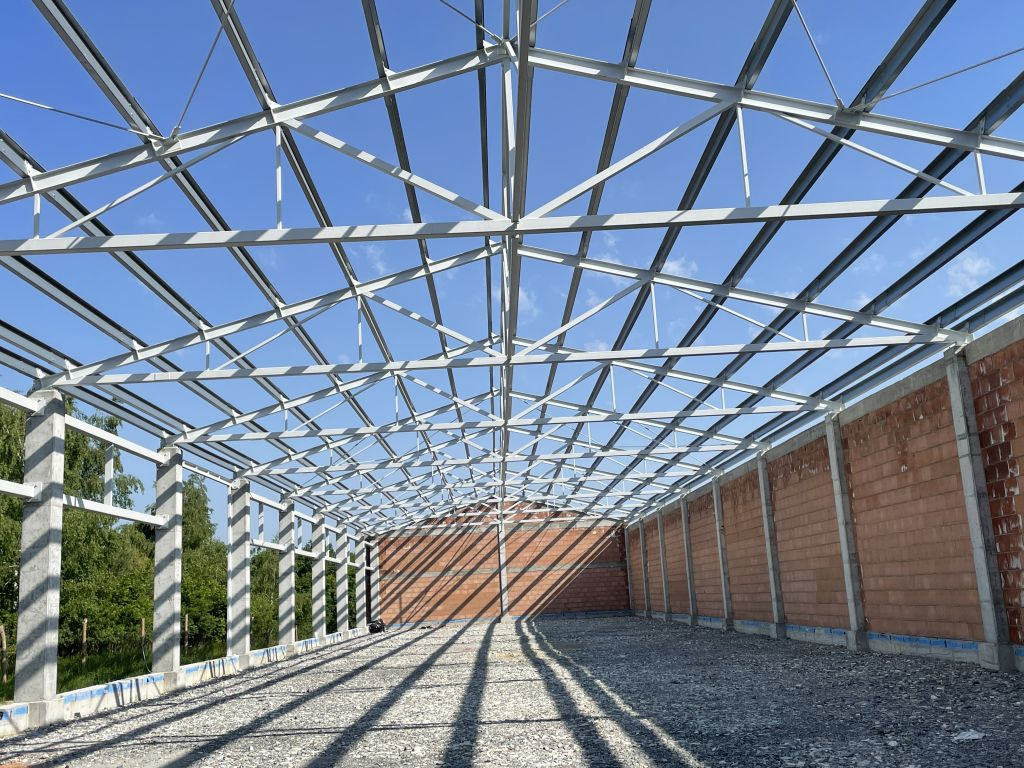 Wnętrze budowanej hali: stalowa konstrukcja szkieletowa, częściowo wymurowane ściany z cegły, żwirowe podłoże, widoczne drzewa w tle, słoneczny dzień.