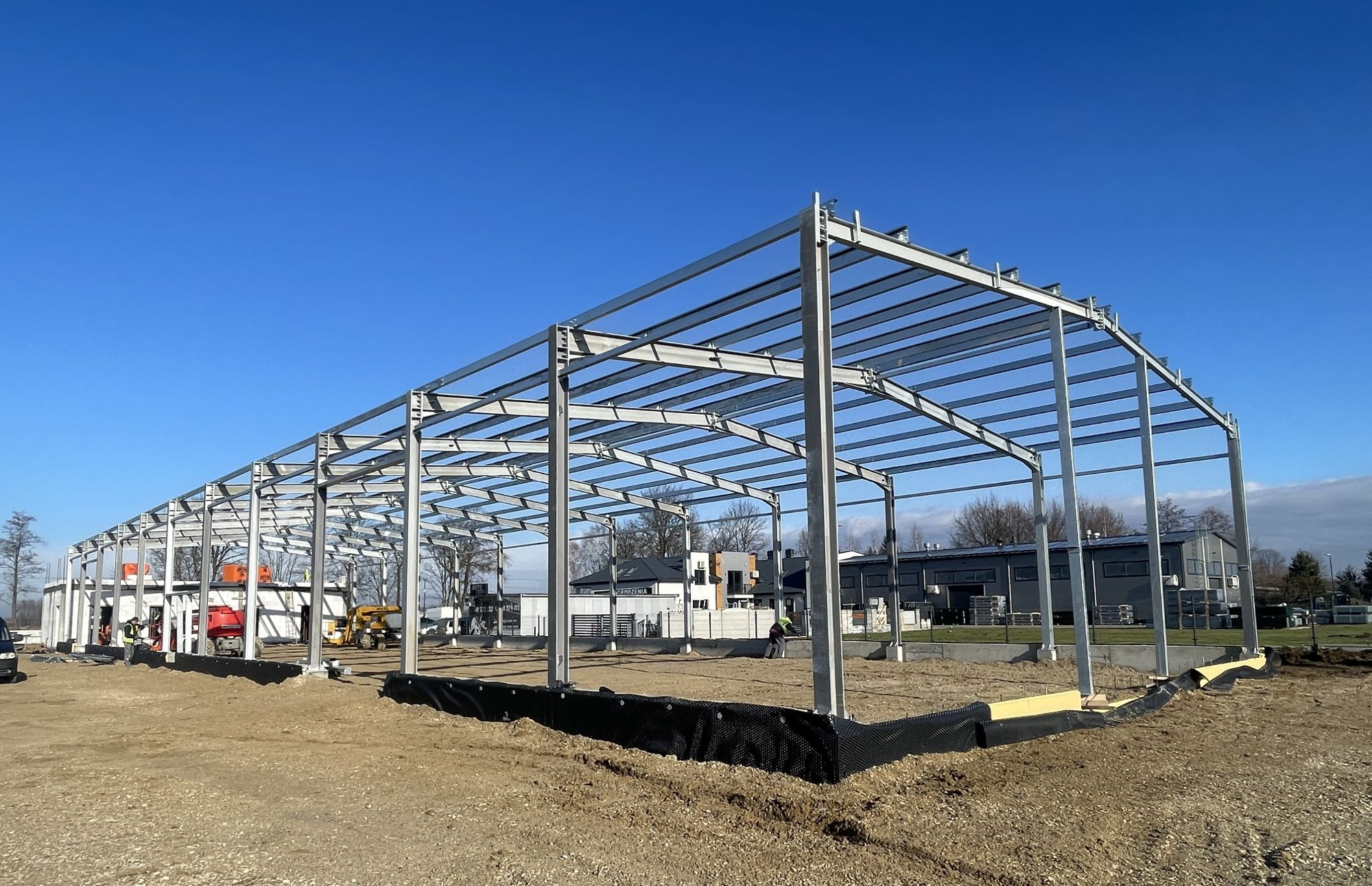 Konstrukcja stalowa hali w trakcie budowy, widok z zewnątrz, błękitne niebo, maszyny budowlane w tle. Widoczne elementy zbrojenia.