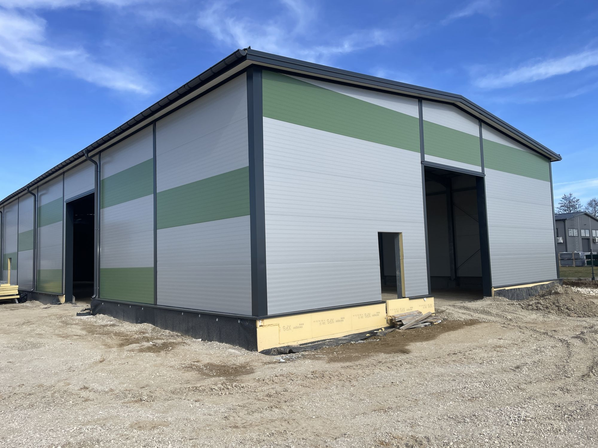 Nowoczesna hala z płyt warstwowych w odcieniach szarości i zieleni, z otwartymi wrotami i widocznym ociepleniem fundamentów, na tle błękitnego nieba.