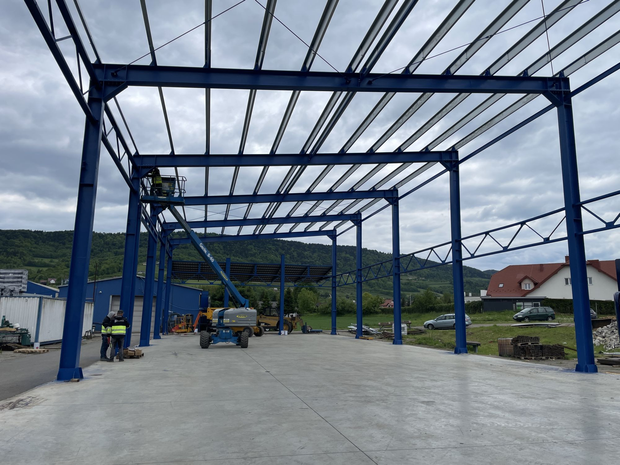 Niebieska konstrukcja hali stalowej w trakcie budowy. Widoczni pracownicy na podnośniku i na ziemi. W tle pagórkowaty krajobraz i budynki.