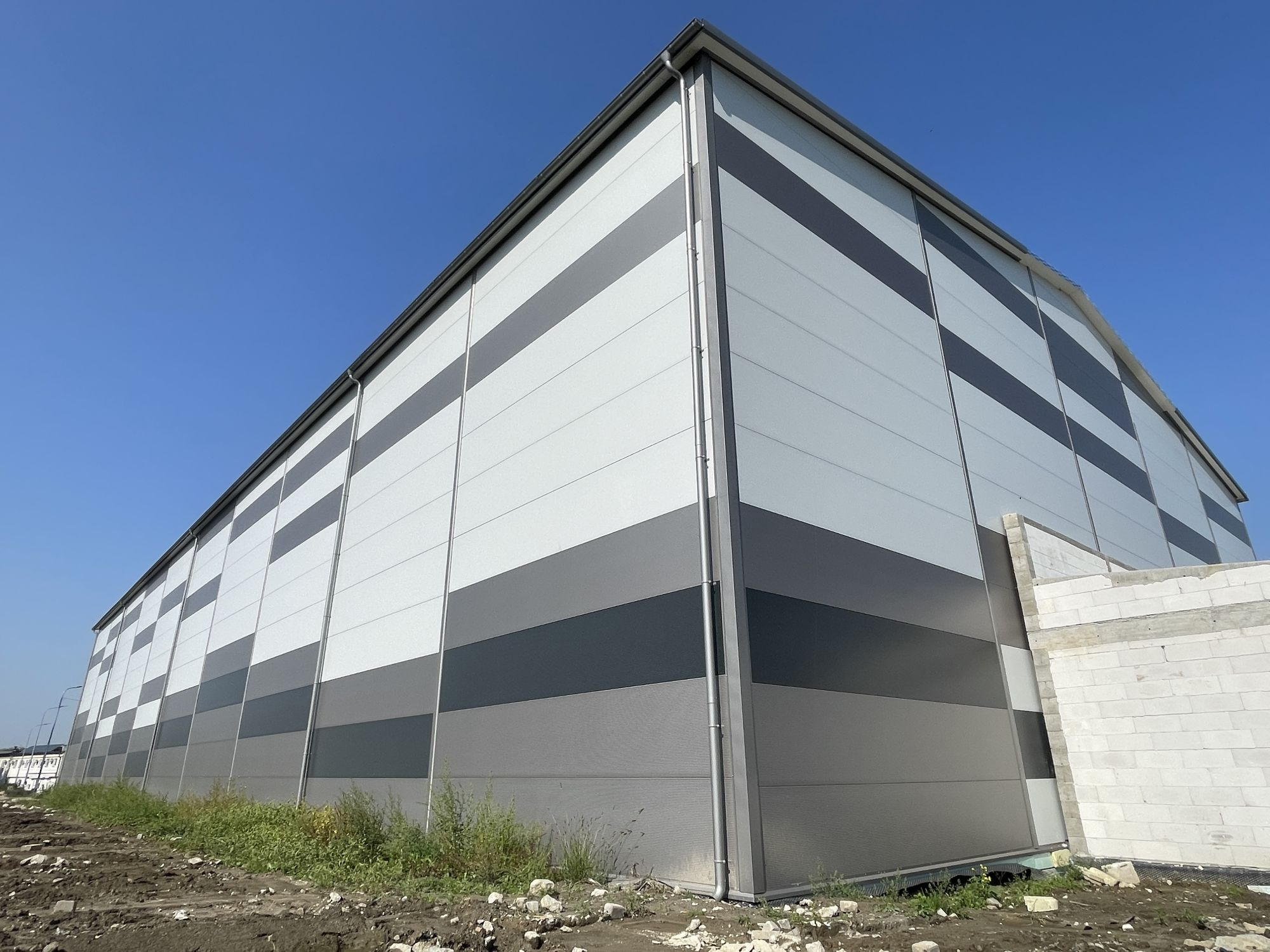 Hala z płyt warstwowych, widok z zewnątrz, z szarymi pasami, błękitne niebo. Częściowo widoczny fundament budynku z białych bloczków.