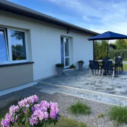 Nowoczesny dom z jasną elewacją, tarasem z kostki brukowej i kwitnącym rododendronem w ogrodzie. Widok na okno z firankami i niebieski parasol.