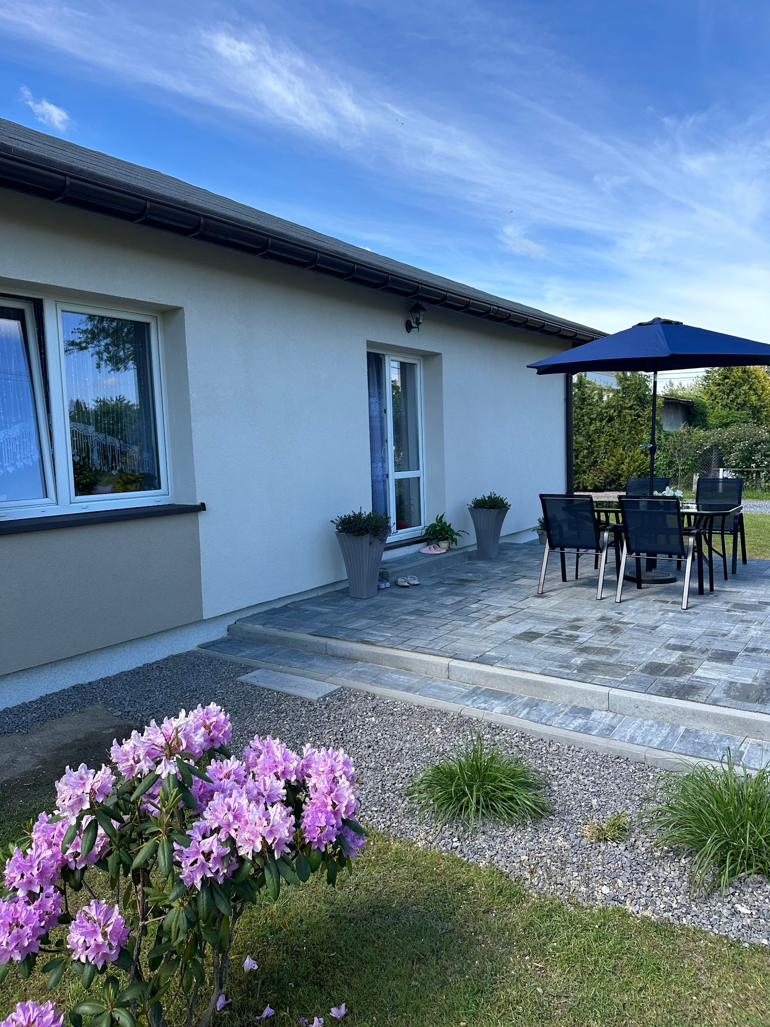 Nowoczesny dom z jasną elewacją, tarasem z kostki brukowej i kwitnącym rododendronem w ogrodzie. Widok na okno z firankami i niebieski parasol.