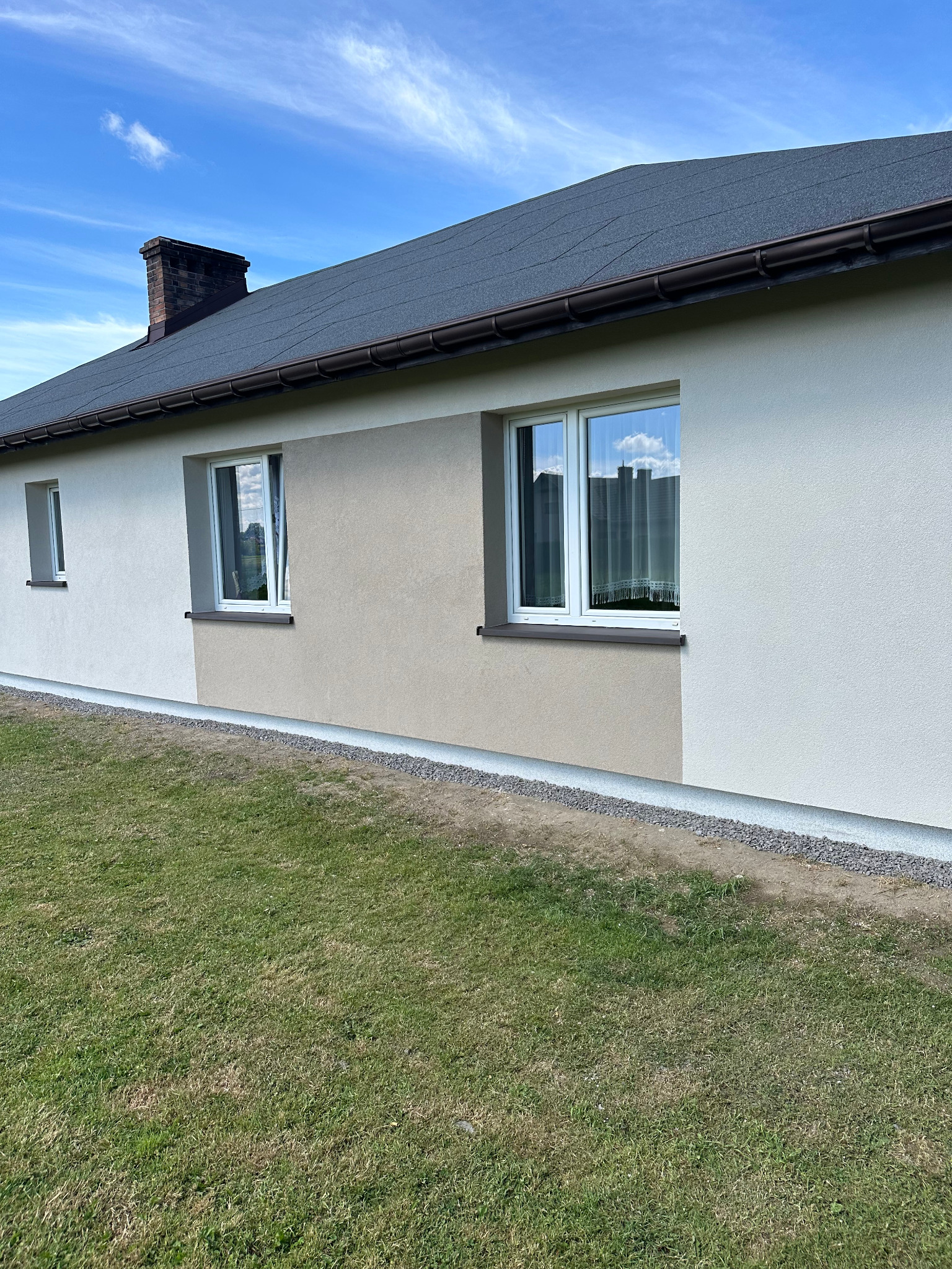 Parterowy dom z dachem pokrytym papą, otynkowany na kremowo i beżowo. Widoczne okna z białymi ramami i ceglany komin. Trawa przed budynkiem. Błękitne niebo.