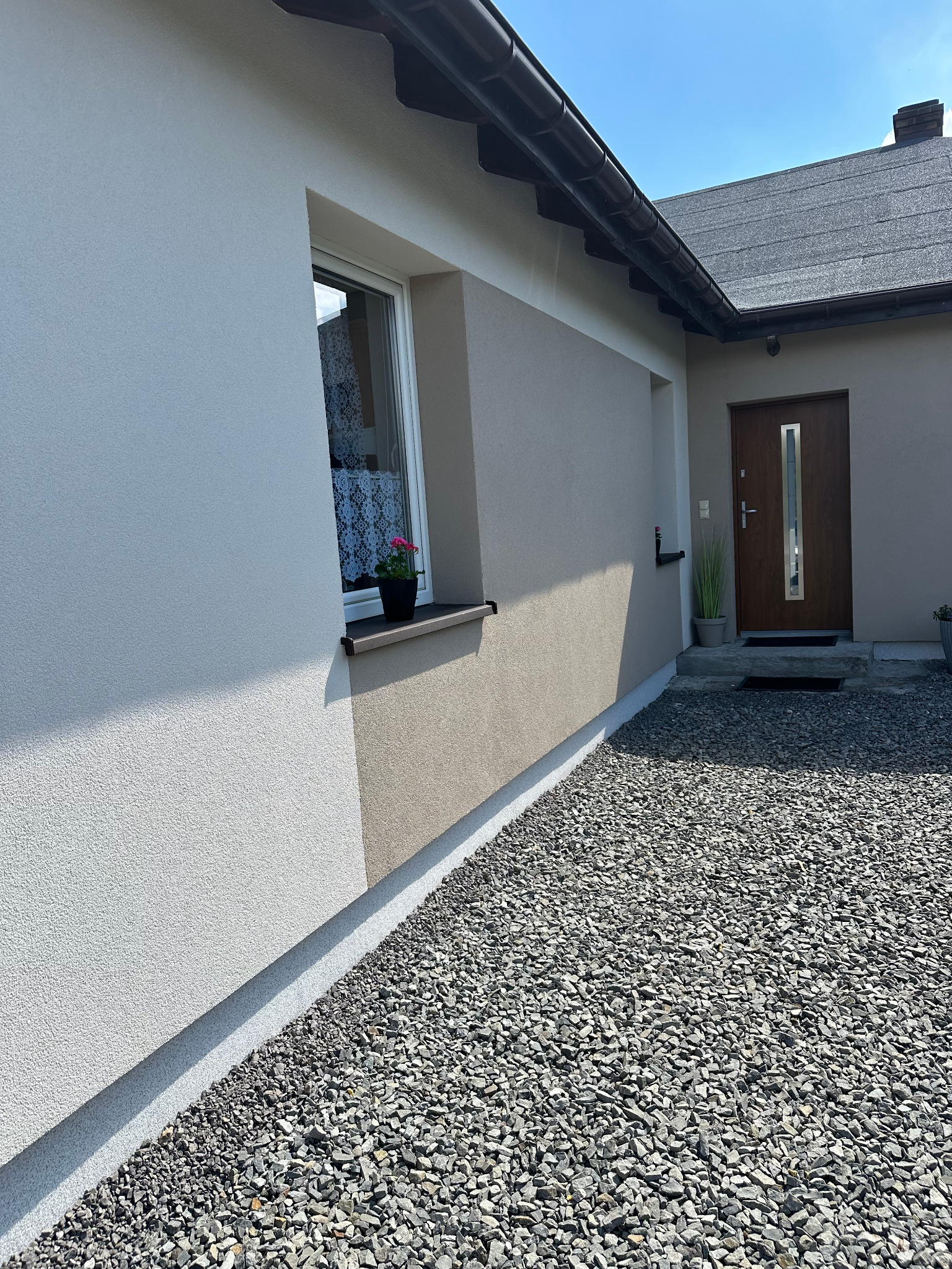 Elewacja domu jednorodzinnego z oknem i kwiatem w doniczce. Widoczne wykończenie w dwóch odcieniach beżu. Podłoże wysypane drobnym kamieniem.