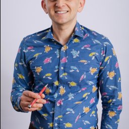 AS SPÓŁKA Z OGRANICZONĄ ODPOWIEDZIALNOŚCIĄ - Uśmiechnięty mężczyzna w koszuli w żółwie, trzymający czerwony długopis, na jasnym tle. Wygląd sugeruje profesjonalizm i otwartość.