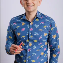 Uśmiechnięty mężczyzna w koszuli w żółwie, trzymający czerwony długopis, na jasnym tle. Wygląd sugeruje profesjonalizm i otwartość.