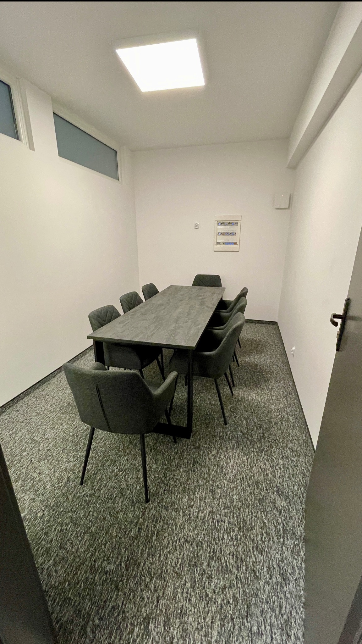 Sala konferencyjna z szarym stołem i krzesłami, oświetlona kwadratową lampą sufitową. Widoczna tablica elektryczna na ścianie i wykładzina dywanowa w odcieniach szarości.