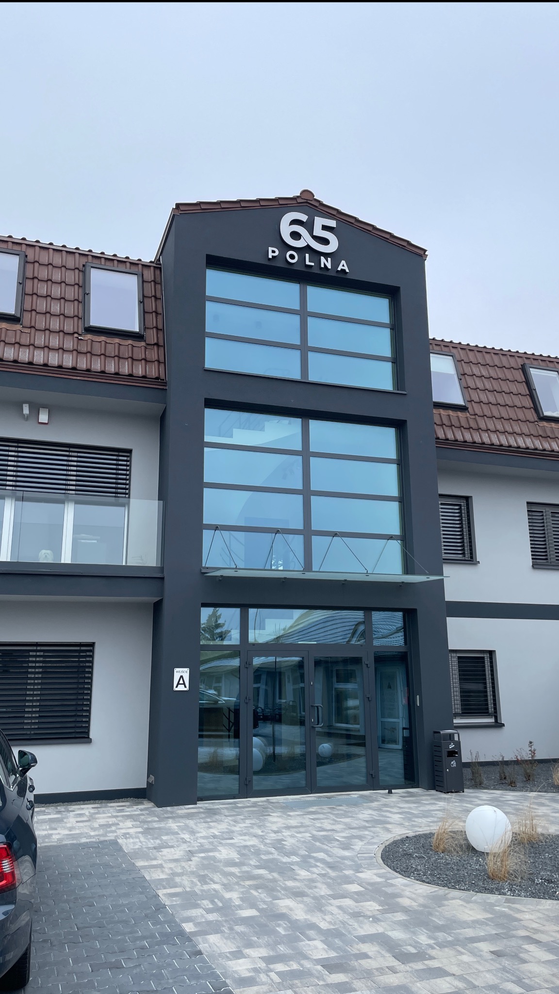 Nowoczesny budynek biurowy z dużymi oknami i numerem '65 POLNA' na szczycie. Szare elewacje, przeszklone drzwi wejściowe i starannie ułożona kostka brukowa na podjeździe.