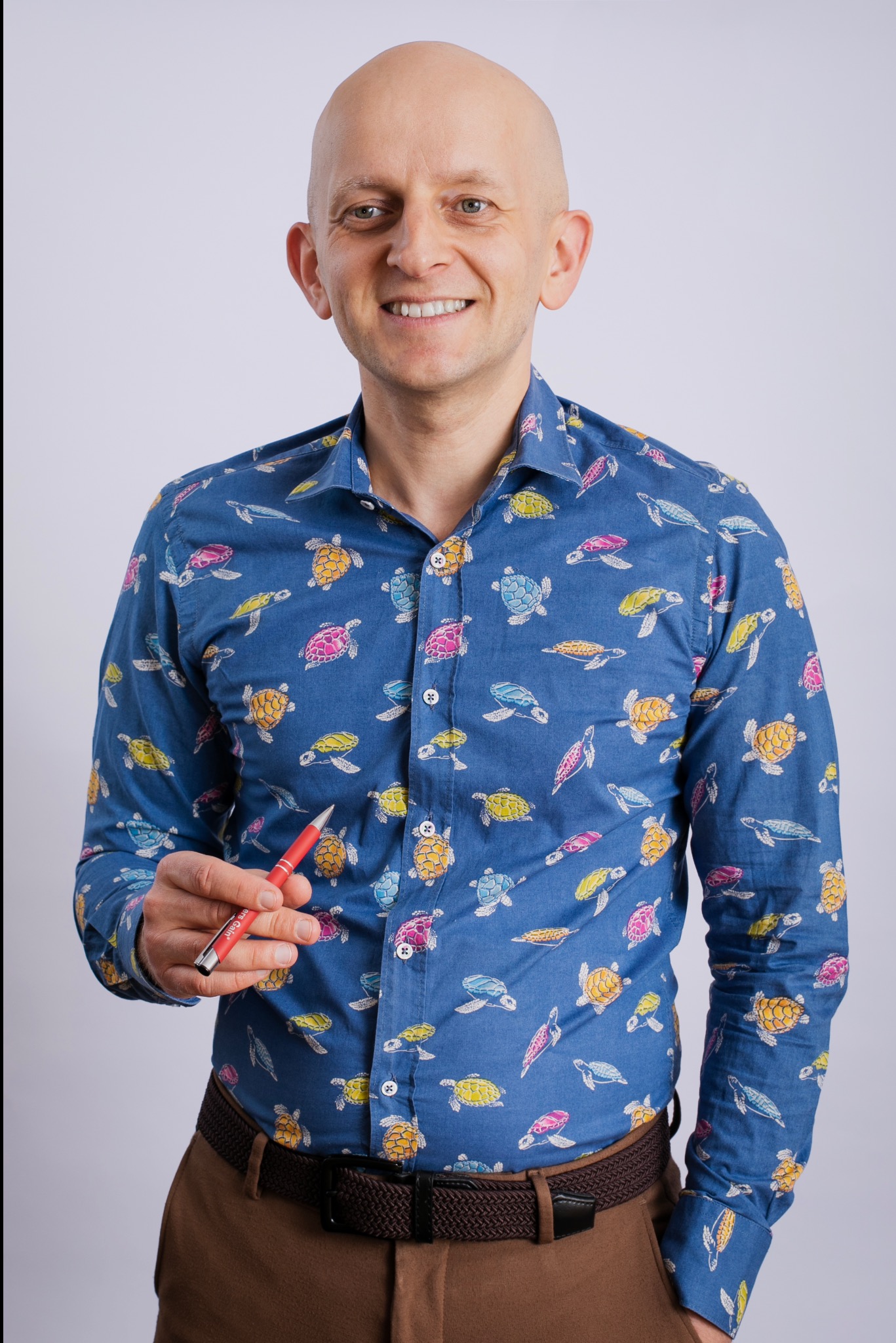 Uśmiechnięty mężczyzna w koszuli w żółwie, trzymający czerwony długopis, na jasnym tle. Wygląd sugeruje profesjonalizm i otwartość.