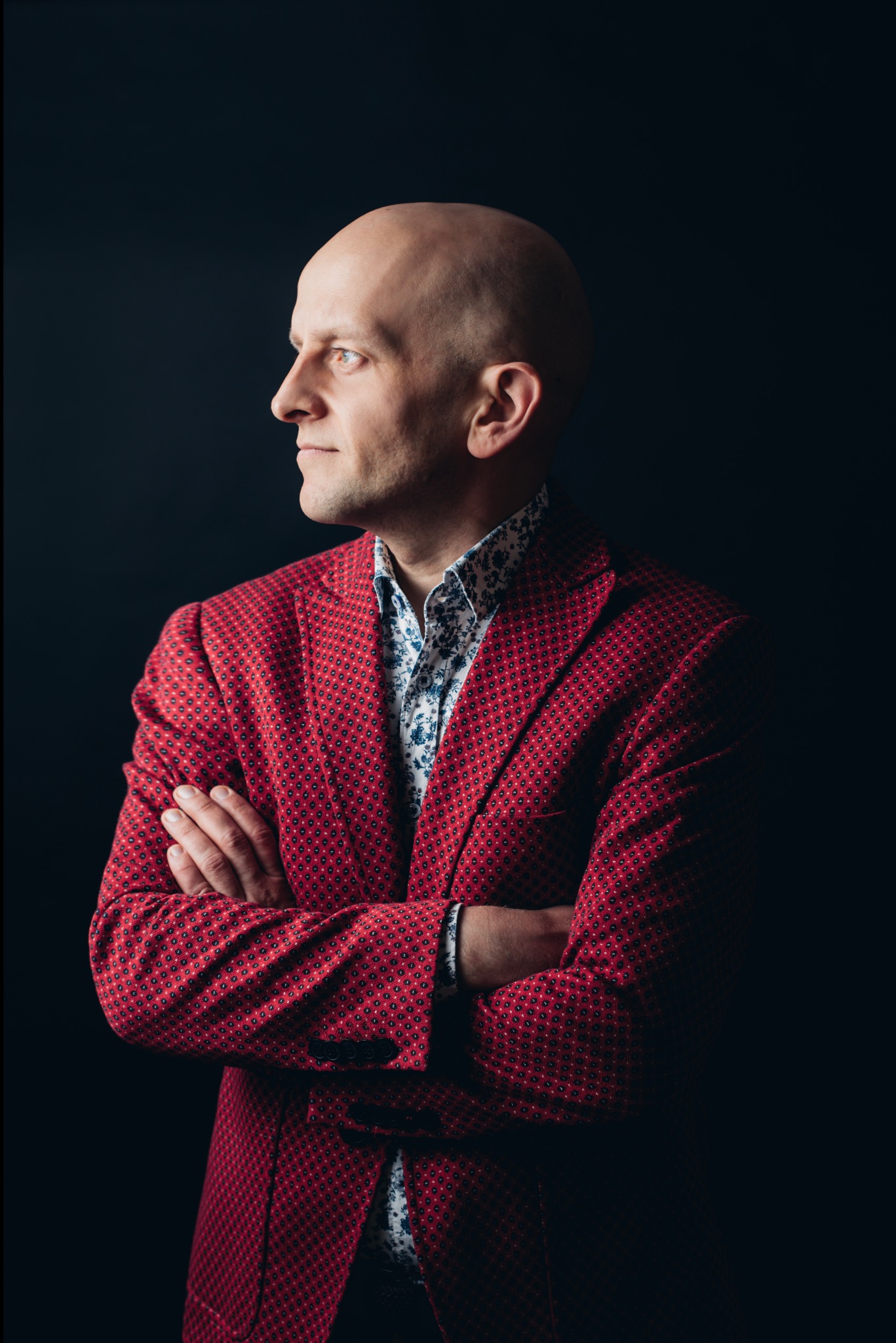 Portret eleganckiego mężczyzny w czerwonej marynarce, z założonymi rękami, patrzy w bok na ciemnym tle. Wyraziste rysy twarzy i wzrok skierowany w dal.