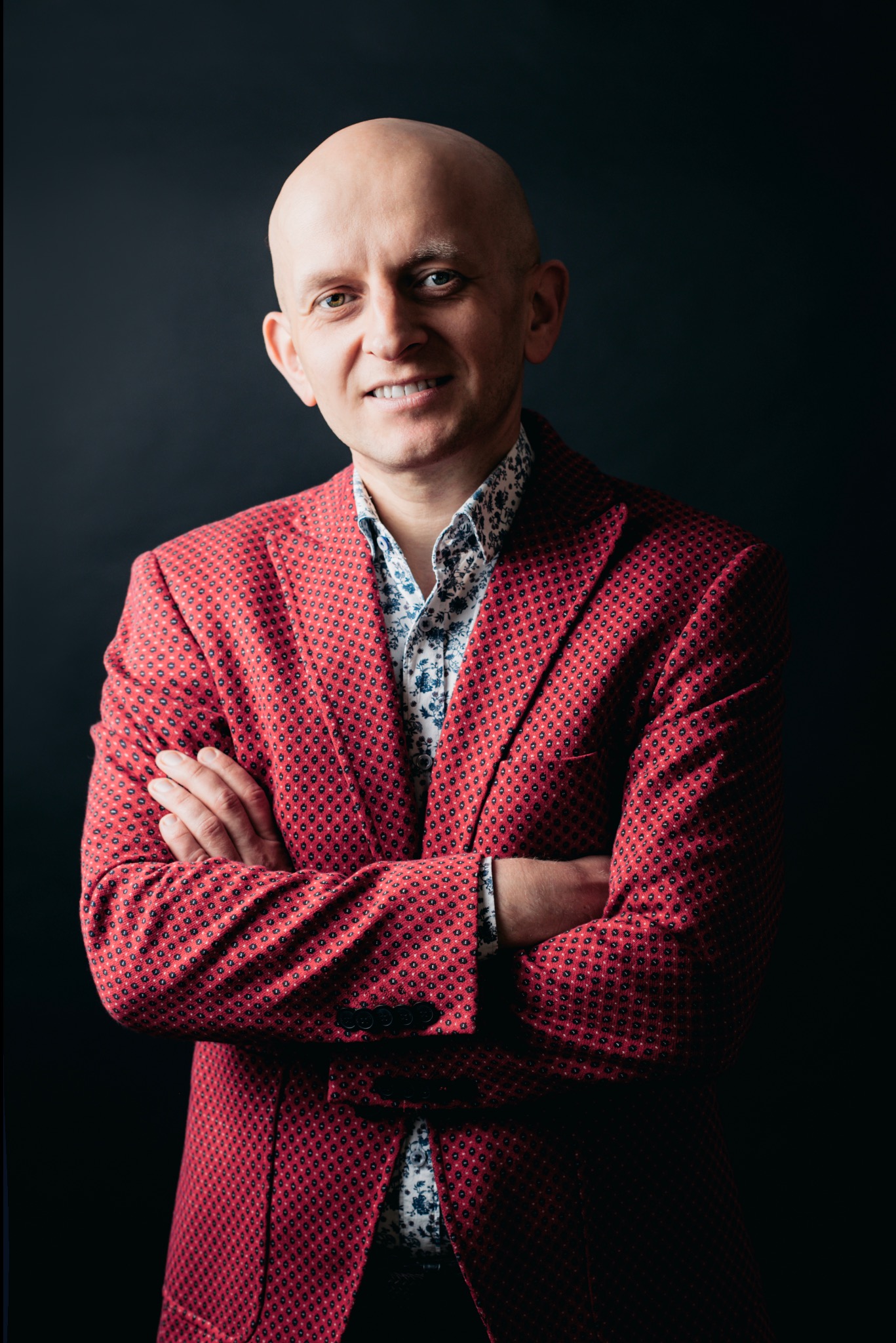 Portret łysiejącego mężczyzny w czerwonej marynarce i wzorzystej koszuli, z założonymi rękami na tle ciemnego studia. Uśmiech, bezpośrednie spojrzenie.