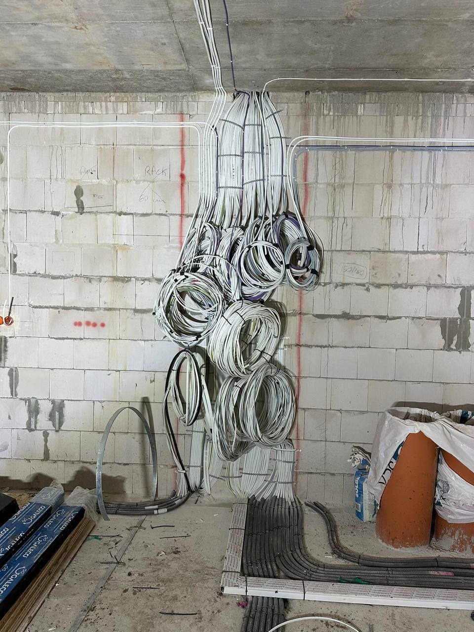 Surowe wnętrze w trakcie prac: wiązki kabli zwisają z sufitu, prowadzone w korytach podłogowych. Widoczne bloczki betonowe i materiały budowlane.