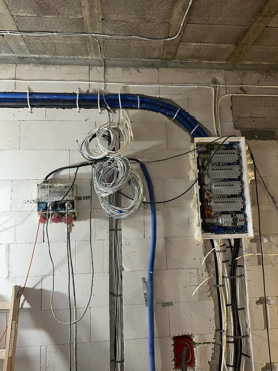 Rozdzielnia elektryczna z niebieskimi peszlami i okablowaniem na surowej ścianie z betonu komórkowego, widoczne zabezpieczenia i przewody. Etap prac.