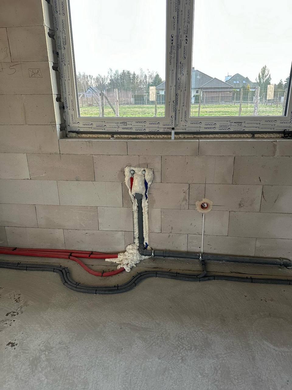 Surowy stan: instalacja elektryczna (peszle) i wodna (rury) w ścianie z betonu komórkowego, okno z widokiem na zewnątrz. Etap prac instalacyjnych w nowym budynku.