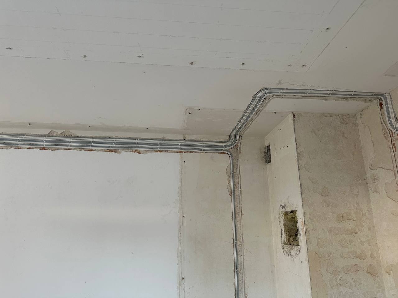 Szare korytka kablowe ułożone wzdłuż ścian i sufitu w surowym wnętrzu. Instalacja elektryczna w trakcie prac wykończeniowych, widoczne ślady po kuciu.