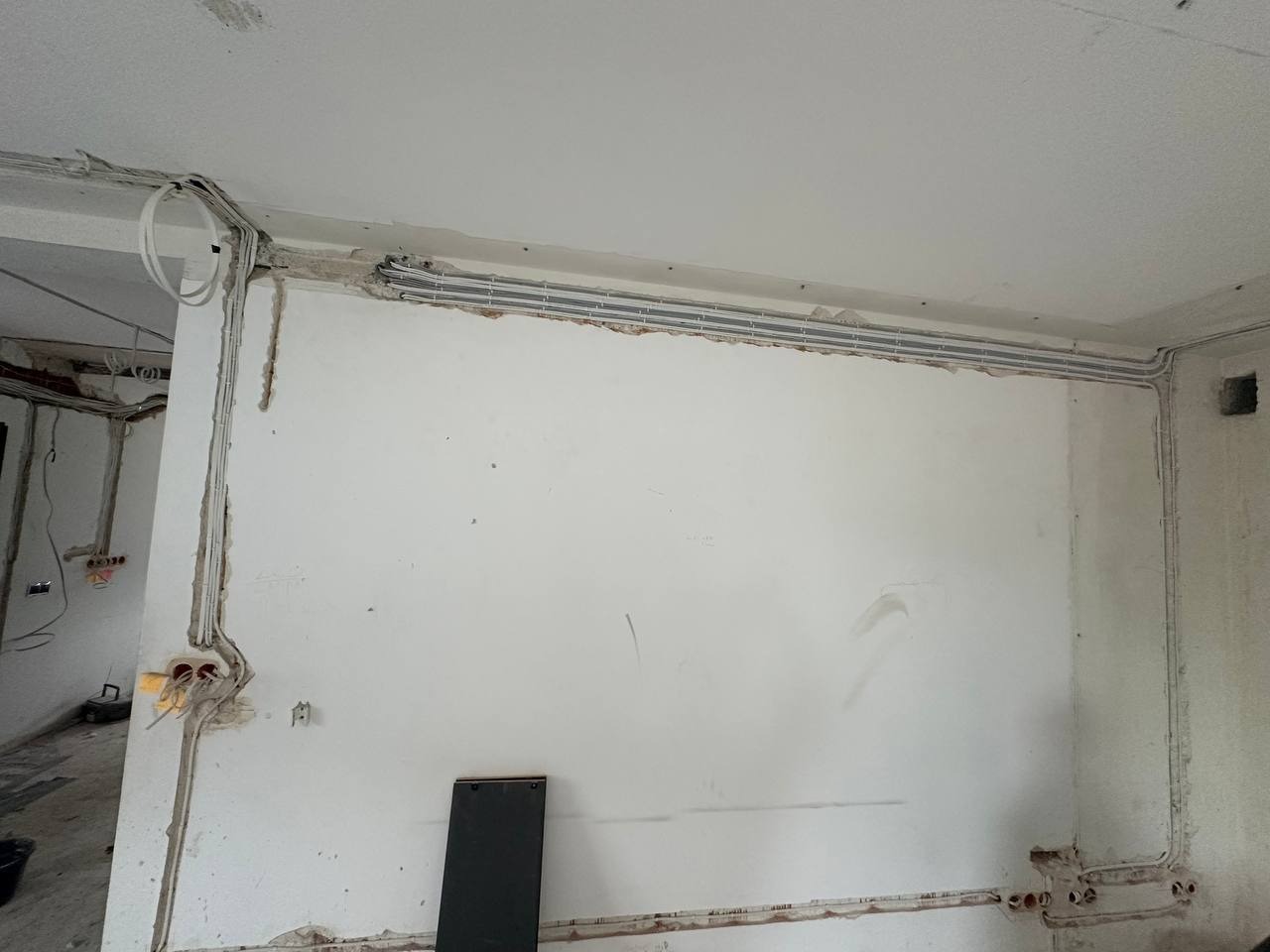 Szare rury instalacji elektrycznej prowadzone pod sufitem i wzdłuż ścian w surowym wnętrzu. Widoczne kable i puszki elektryczne w ścianach. Etap prac instalacyjnych.