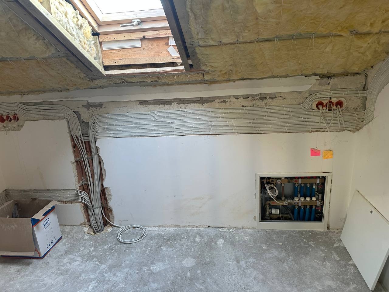 Rozprowadzone kable elektryczne i rury w surowym wnętrzu poddasza z oknem dachowym, widoczna instalacja wodna w ścianie. Elementy izolacji na stropie.