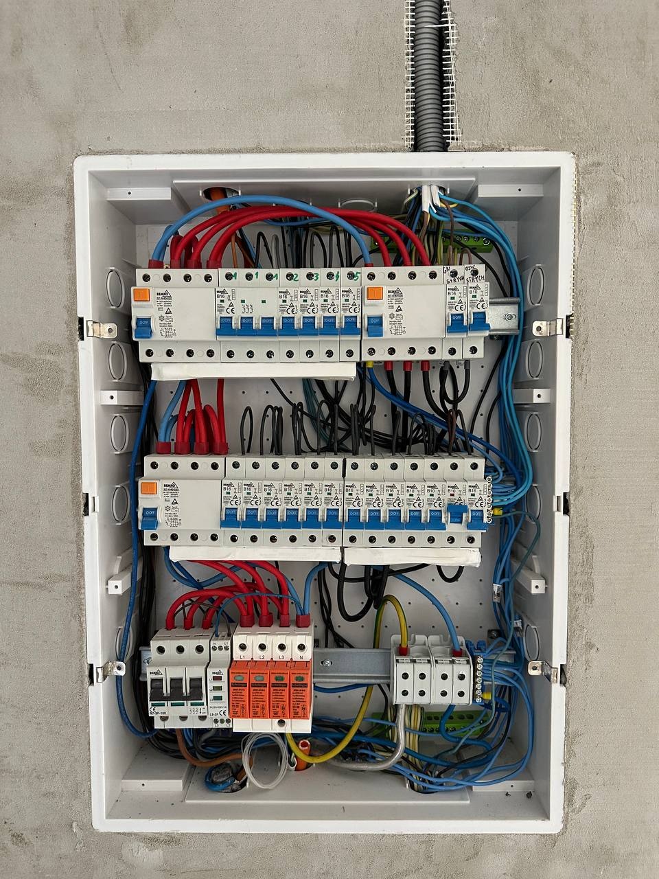 Rozdzielnia elektryczna w białej obudowie, z widocznymi bezpiecznikami, przewodami w kolorach niebieskim i czerwonym, oraz elementami instalacji odgromowej.
