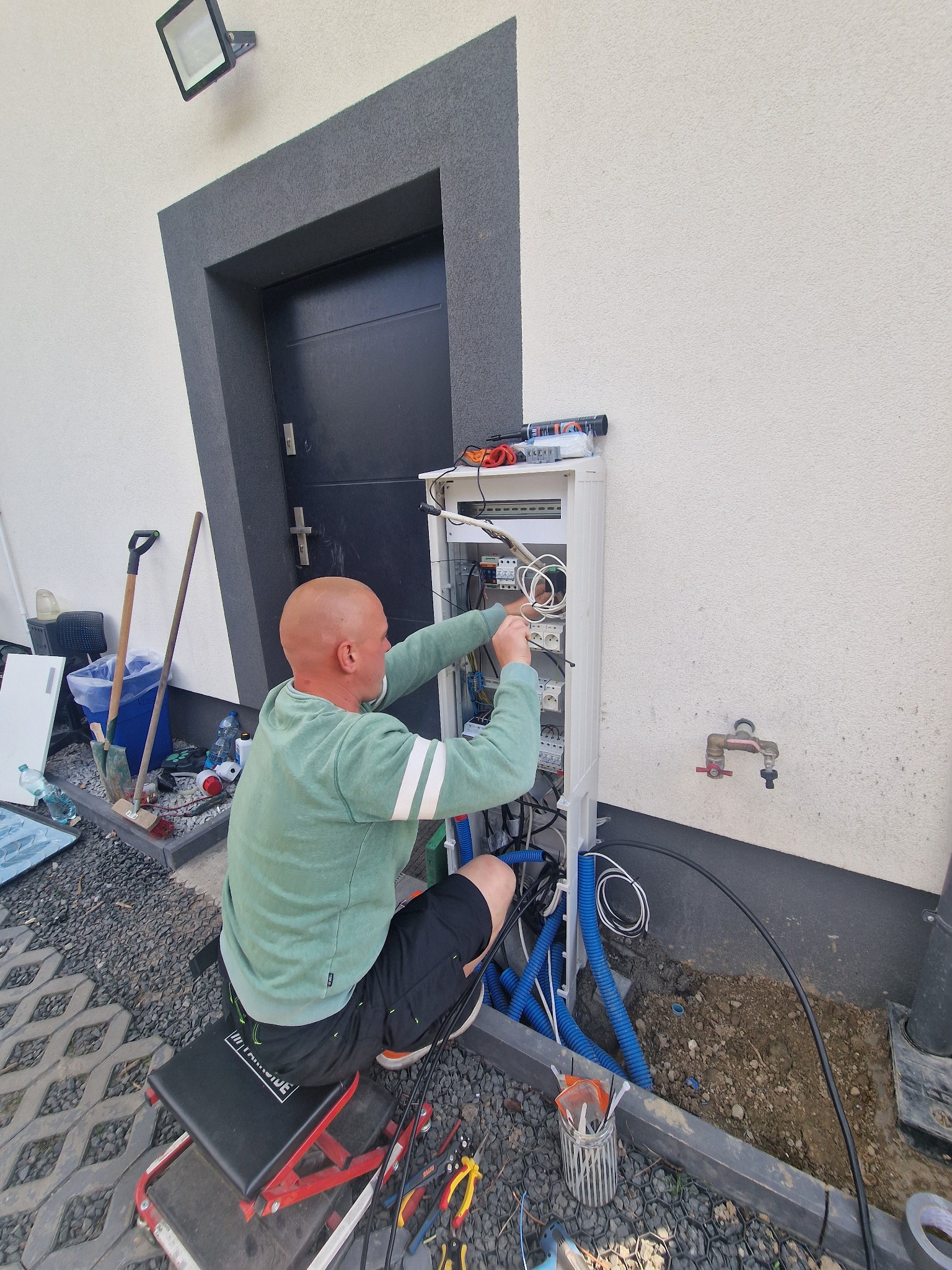 Elektryk w trakcie podłączania instalacji elektrycznej w białej skrzynce na elewacji domu. Widoczne narzędzia i przewody.