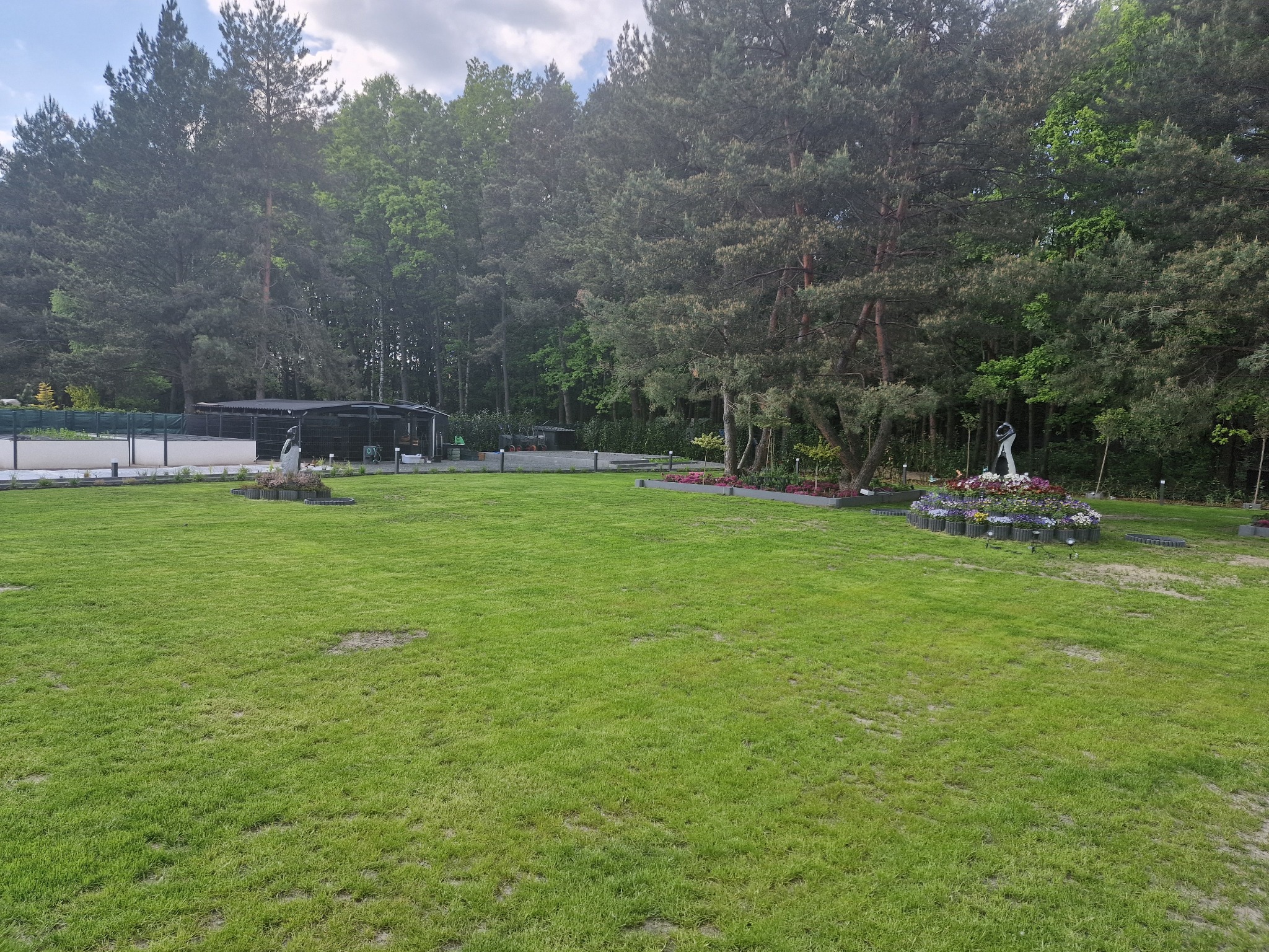 Rozległy, zielony trawnik z rzeźbami i kwiatami, otoczony drzewami. Widoczne elementy małej architektury ogrodowej i oświetlenie. Całość utrzymana w estetycznym porządku.