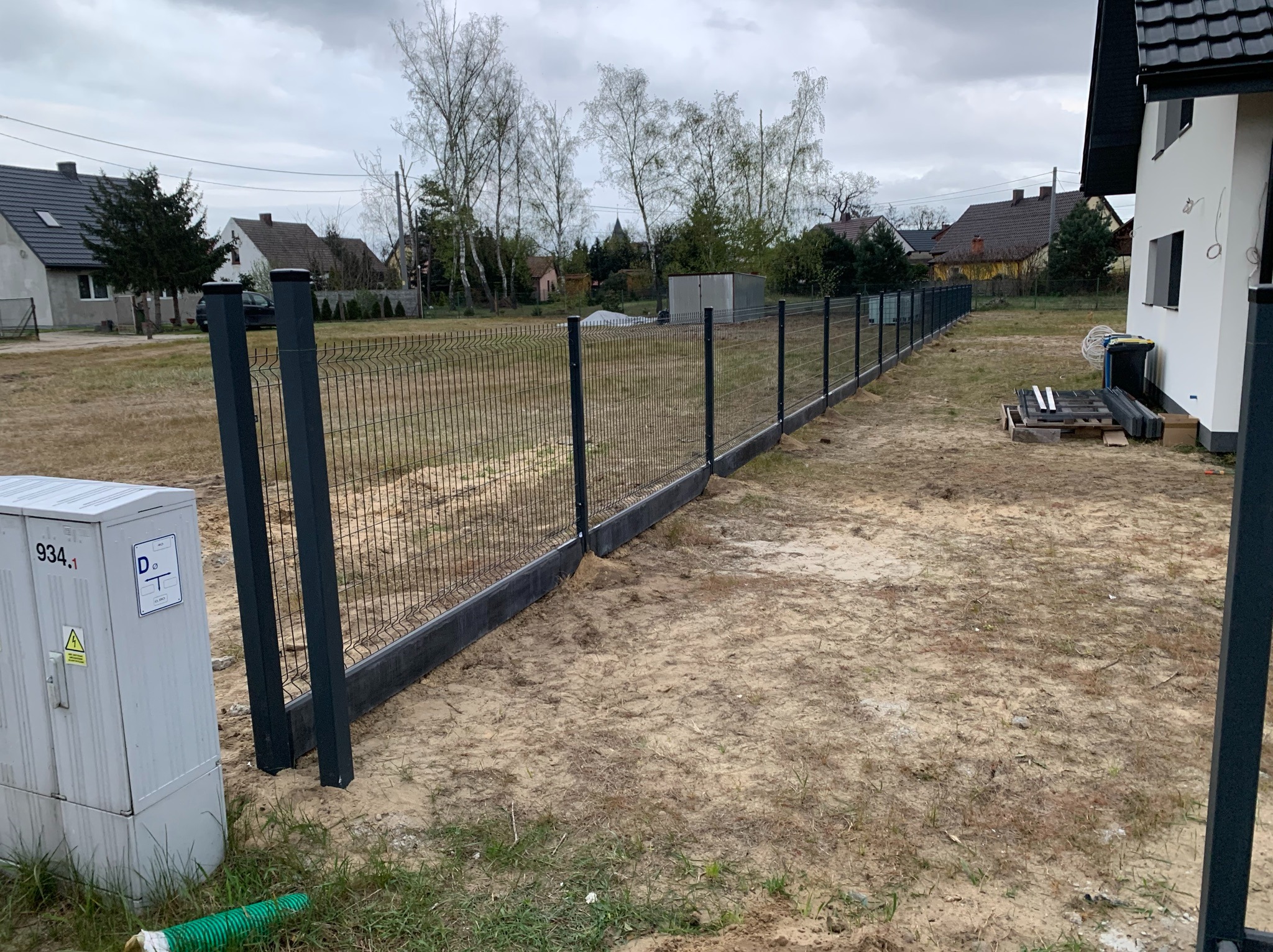Nowoczesne ogrodzenie panelowe z podmurówką betonową, widoczne z perspektywy bocznej, na tle domów jednorodzinnych w pochmurny dzień. Elementy montażowe obok budynku.