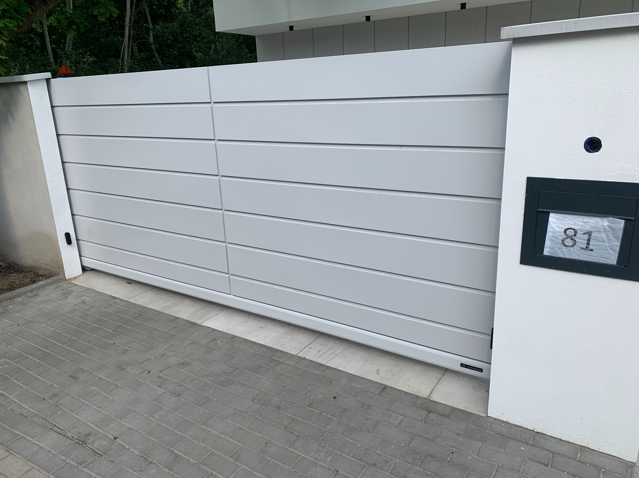 Nowoczesna, przesuwna brama wjazdowa w odcieniach szarości, z numerem domu '81' na białym słupku. Widoczny fragment kostki brukowej i zieleni w tle.