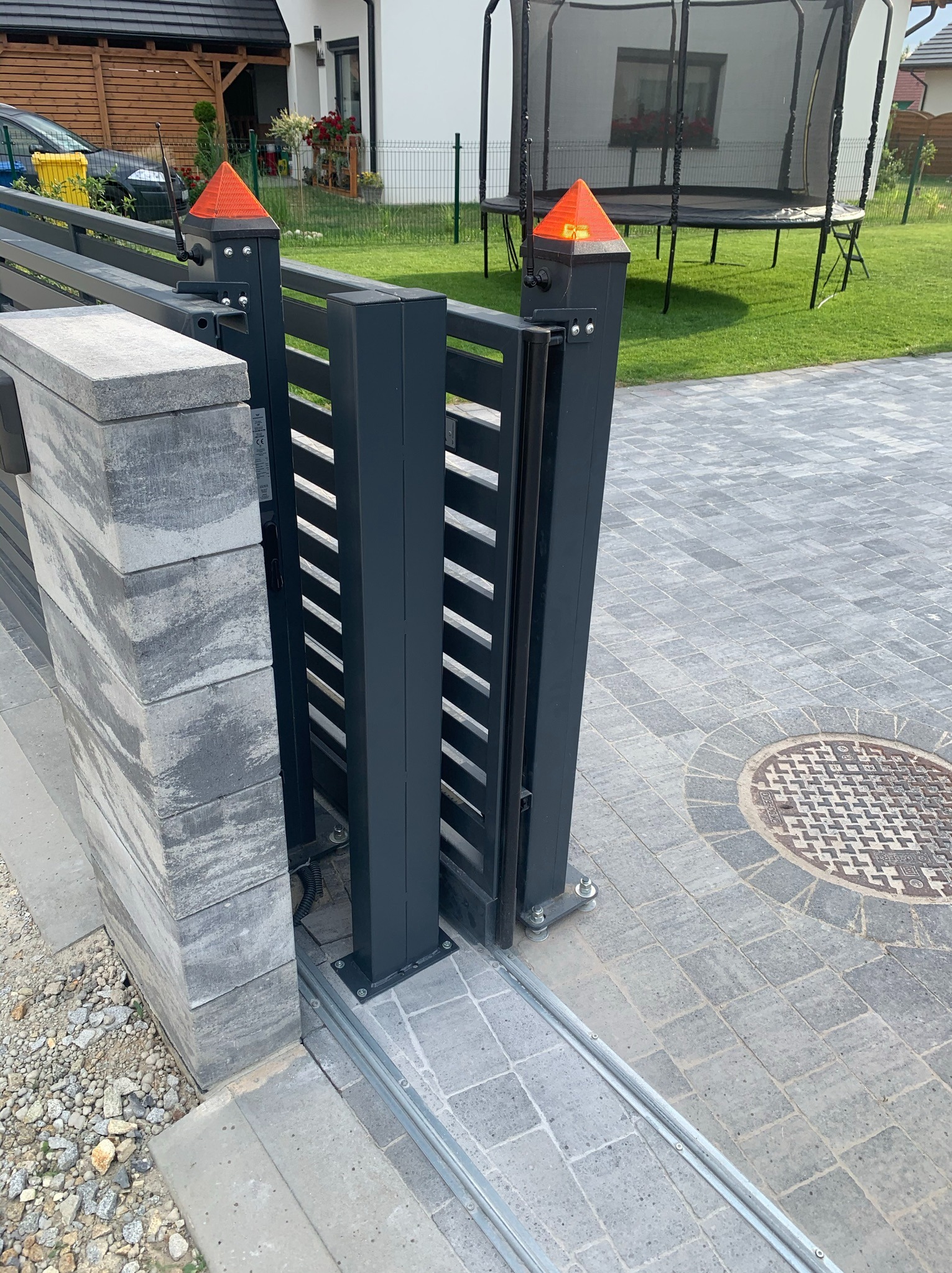 Nowoczesna, przesuwna brama wjazdowa z automatyką, częściowo otwarta. Widoczna szyna, słupki z pomarańczowymi lampkami ostrzegawczymi i kostka brukowa. Brama w kolorze grafitowym.