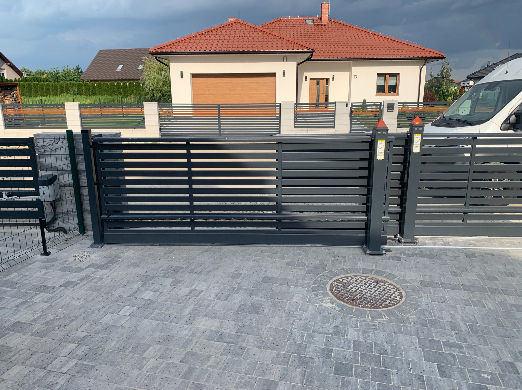 Nowoczesna, przesuwna brama wjazdowa w kolorze grafitowym, z poziomymi panelami, na tle domu z czerwoną dachówką i szarej kostki brukowej. Widoczny fragment ogrodzenia.