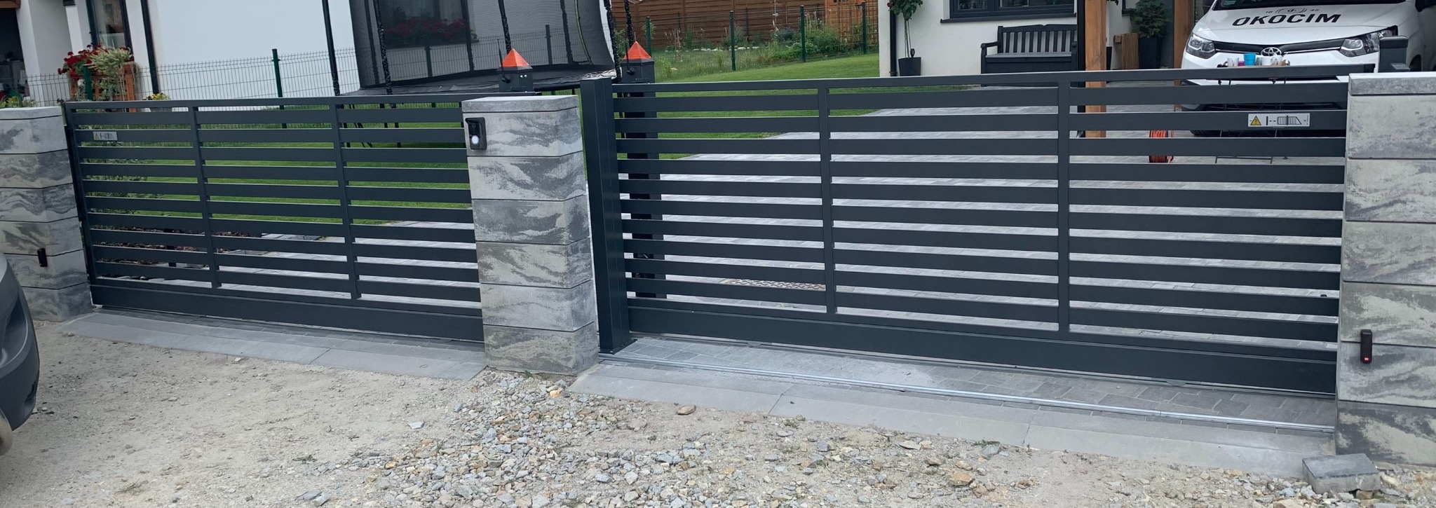 Nowoczesna brama przesuwna z poziomymi panelami w kolorze grafitowym, osadzona na słupkach z szarego kamienia. Widoczny dom i zaparkowany samochód w tle.