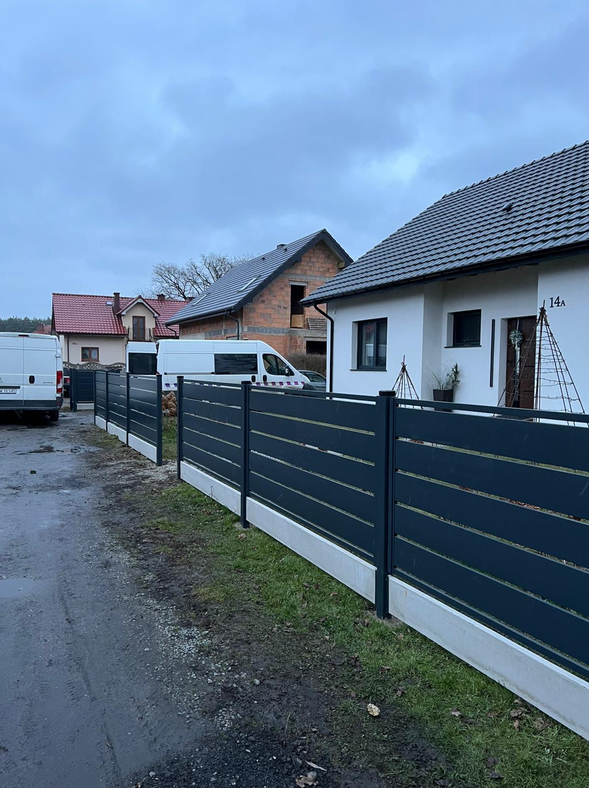 Nowoczesne, poziome ogrodzenie panelowe w kolorze grafitowym z betonową podmurówką, na tle domów jednorodzinnych i zaparkowanych busów. Pochmurne niebo.