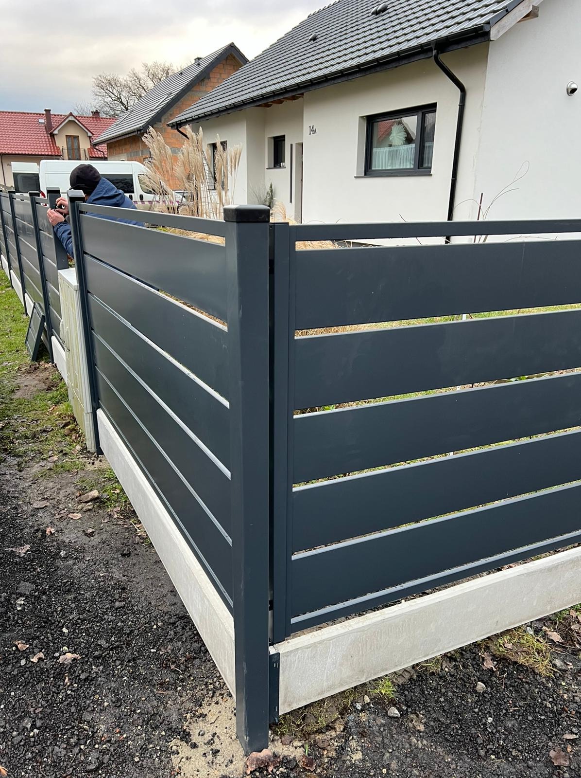 Nowoczesne, poziome ogrodzenie metalowe w kolorze grafitowym, z widocznym montażem przez montera. Tło: dom z białymi ścianami i szarą dachówką.