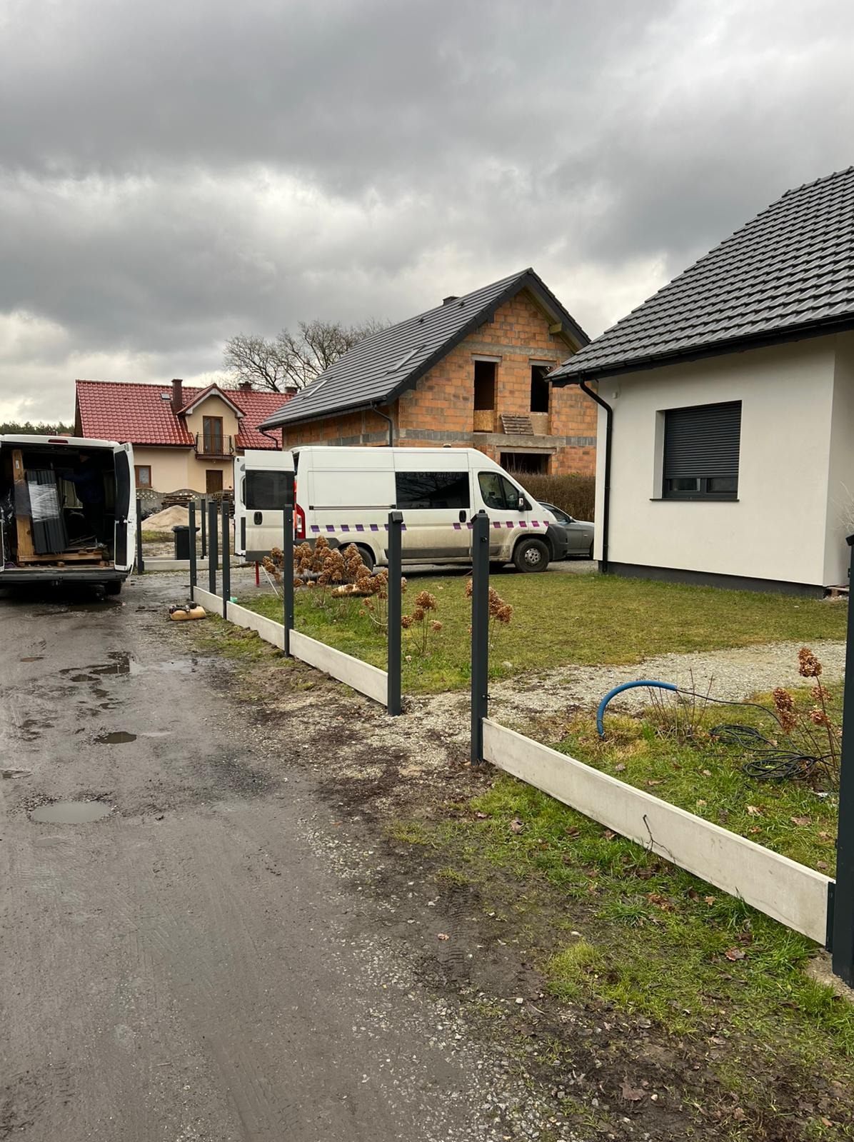 Montaż ogrodzenia panelowego: betonowa podmurówka i słupki w kolorze grafitowym, na tle dwóch dostawczych aut i budynków w różnym stanie wykończenia, pochmurny dzień.