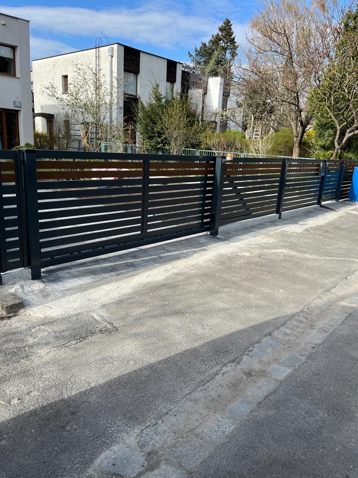 Nowoczesne ogrodzenie posesji z poziomych, ciemnych paneli metalowych i drewnianych akcentów, na tle zabudowy jednorodzinnej w słoneczny dzień.