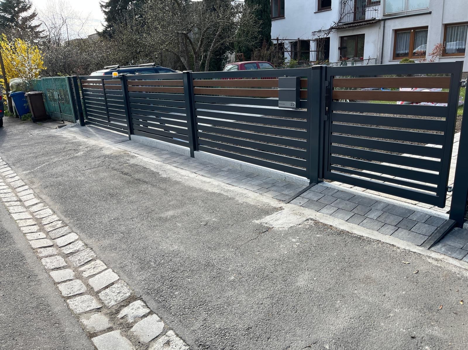 Nowoczesna brama wjazdowa z poziomymi, szarymi przęsłami i drewnianymi akcentami, skrzynka na listy w kolorze bramy, kostka brukowa, dom w tle.