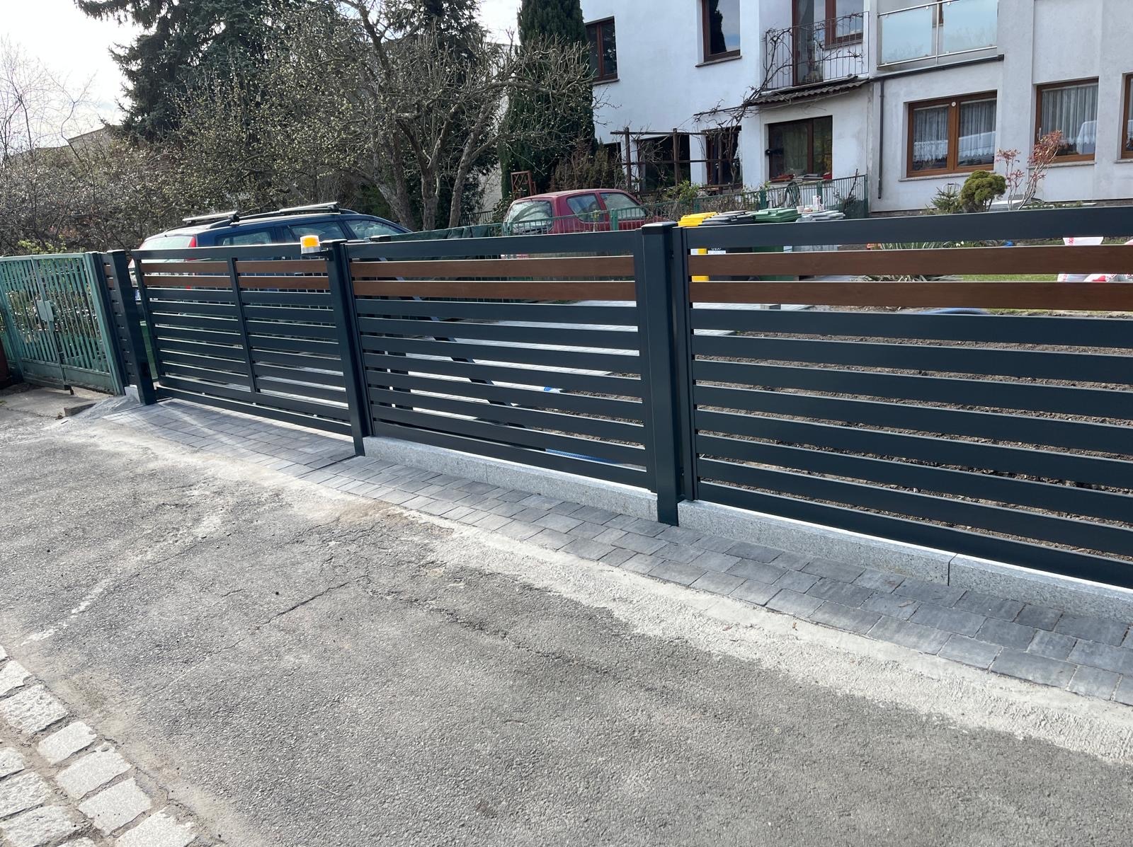 Nowoczesna brama wjazdowa z poziomymi, grafitowymi przęsłami i drewnianym akcentem u góry, osadzona na kostce brukowej. W tle budynek mieszkalny i zieleń.
