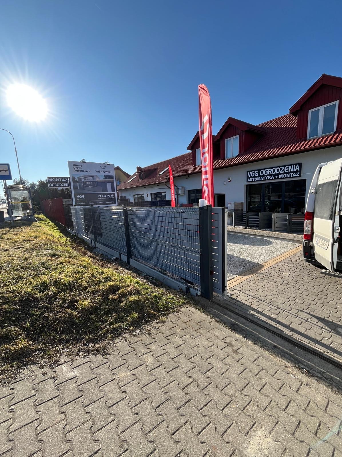 Szara brama przesuwna z wypełnieniem panelowym na tle budynku firmy VGG Ogrodzenia i reklamy Wiśniowski. Widoczne czerwone flagi reklamowe i fragment chodnika z kostki.