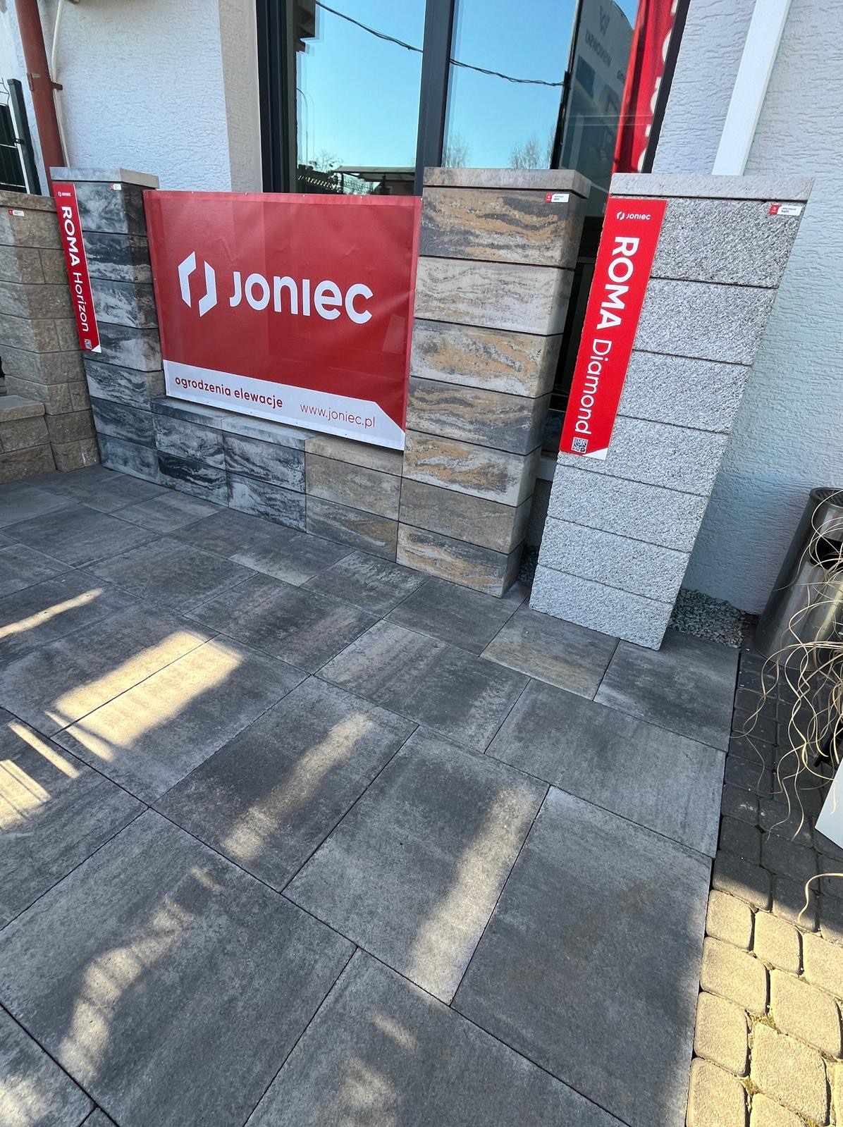 Prezentacja kostki brukowej Joniec Roma Horizon i Diamond, ułożonej na powierzchni, z ekspozycją elementów ogrodzeniowych w tle. Widoczne detale faktury i koloru.