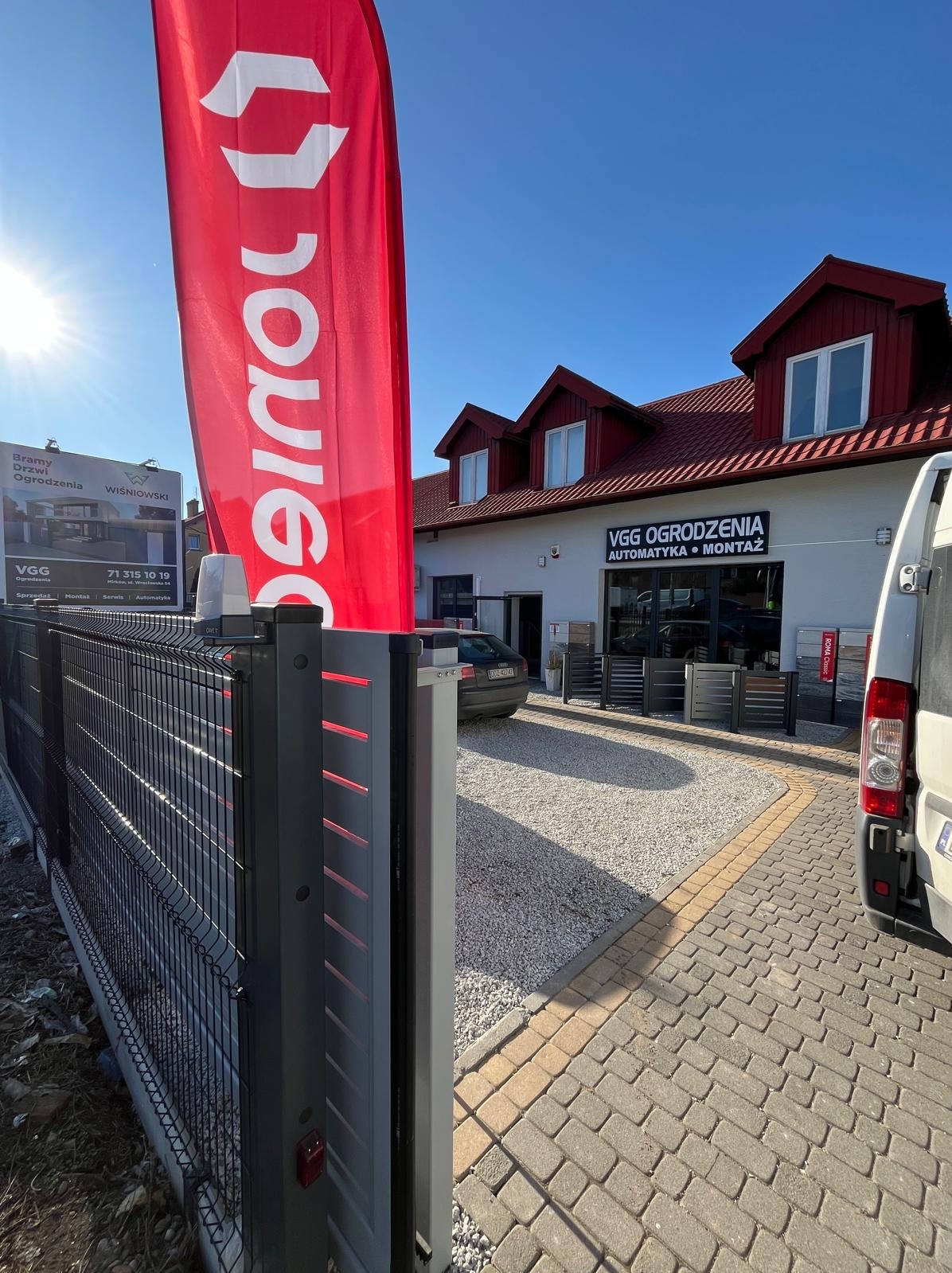 Nowoczesne ogrodzenie panelowe z oświetleniem LED, widok z zewnątrz, na tle siedziby firmy VGG Ogrodzenia z flagą reklamową. Słoneczny dzień.