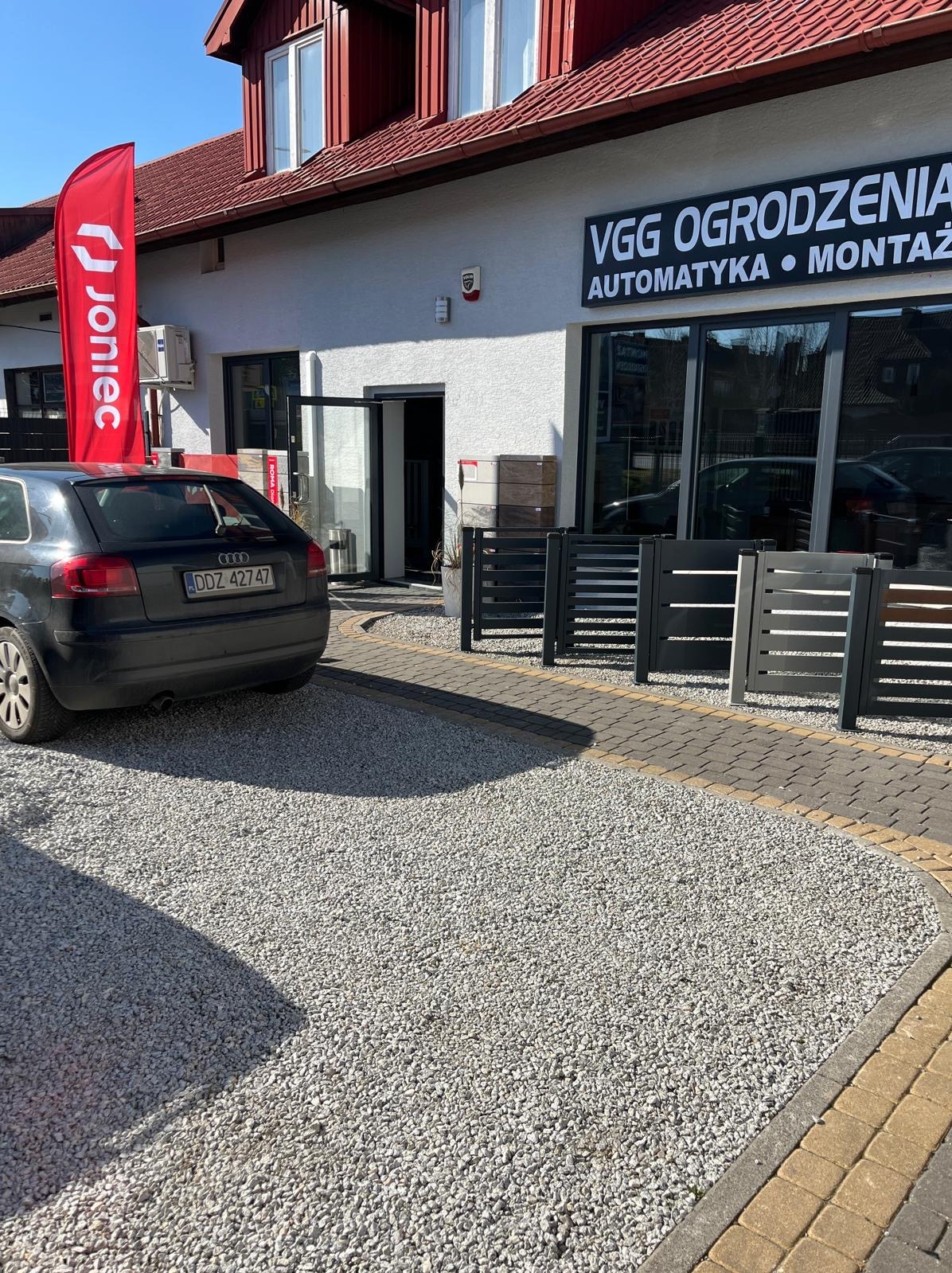 Ekspozycja paneli ogrodzeniowych przed budynkiem firmy VGG Ogrodzenia z automatyką i montażem, widoczny samochód i flaga reklamowa Joniec.