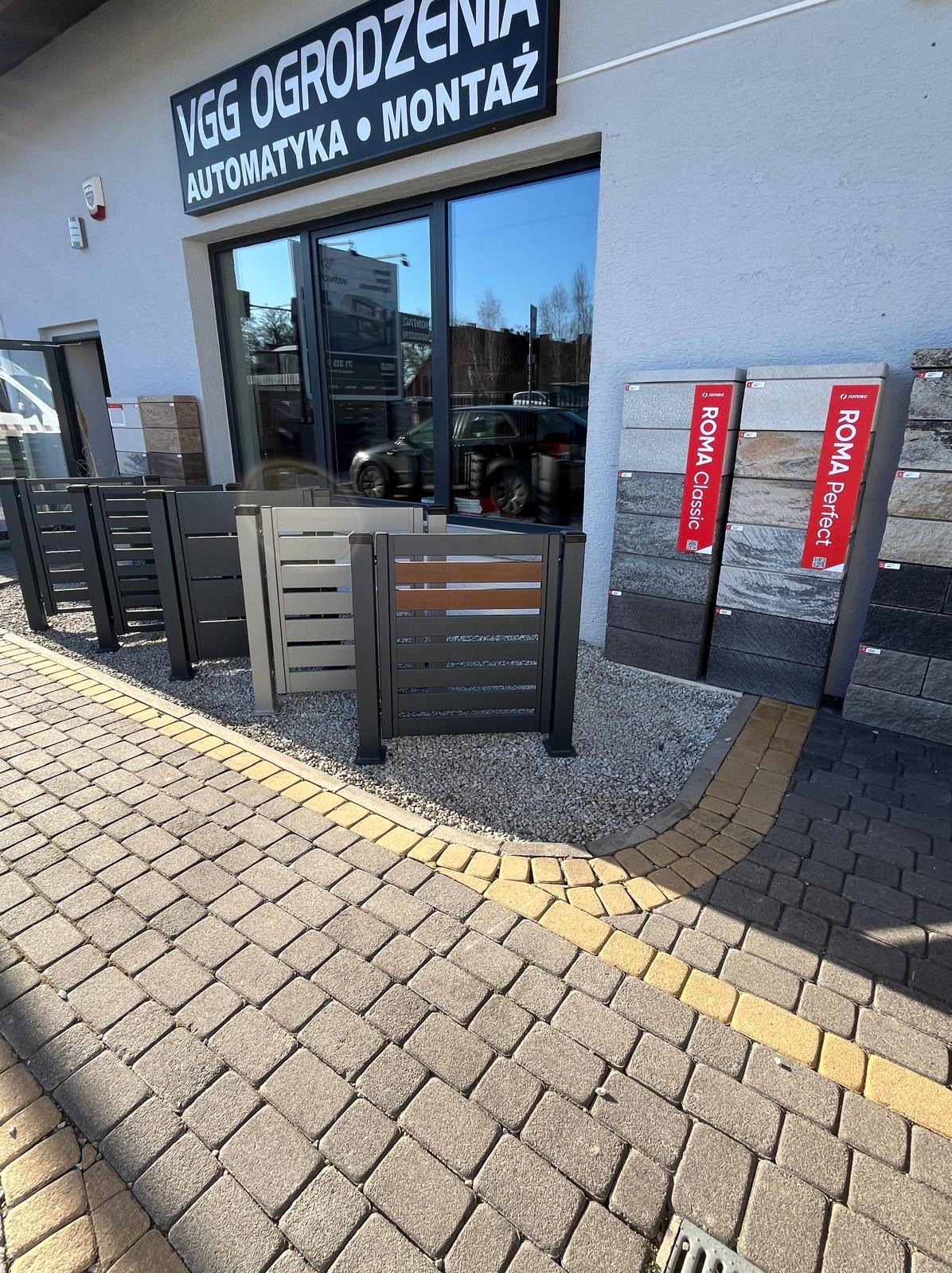 Ekspozycja paneli ogrodzeniowych w różnych kolorach i wzorach przed salonem VGG Ogrodzenia. Widoczne próbki kostki brukowej Roma Classic i Roma Perfect.