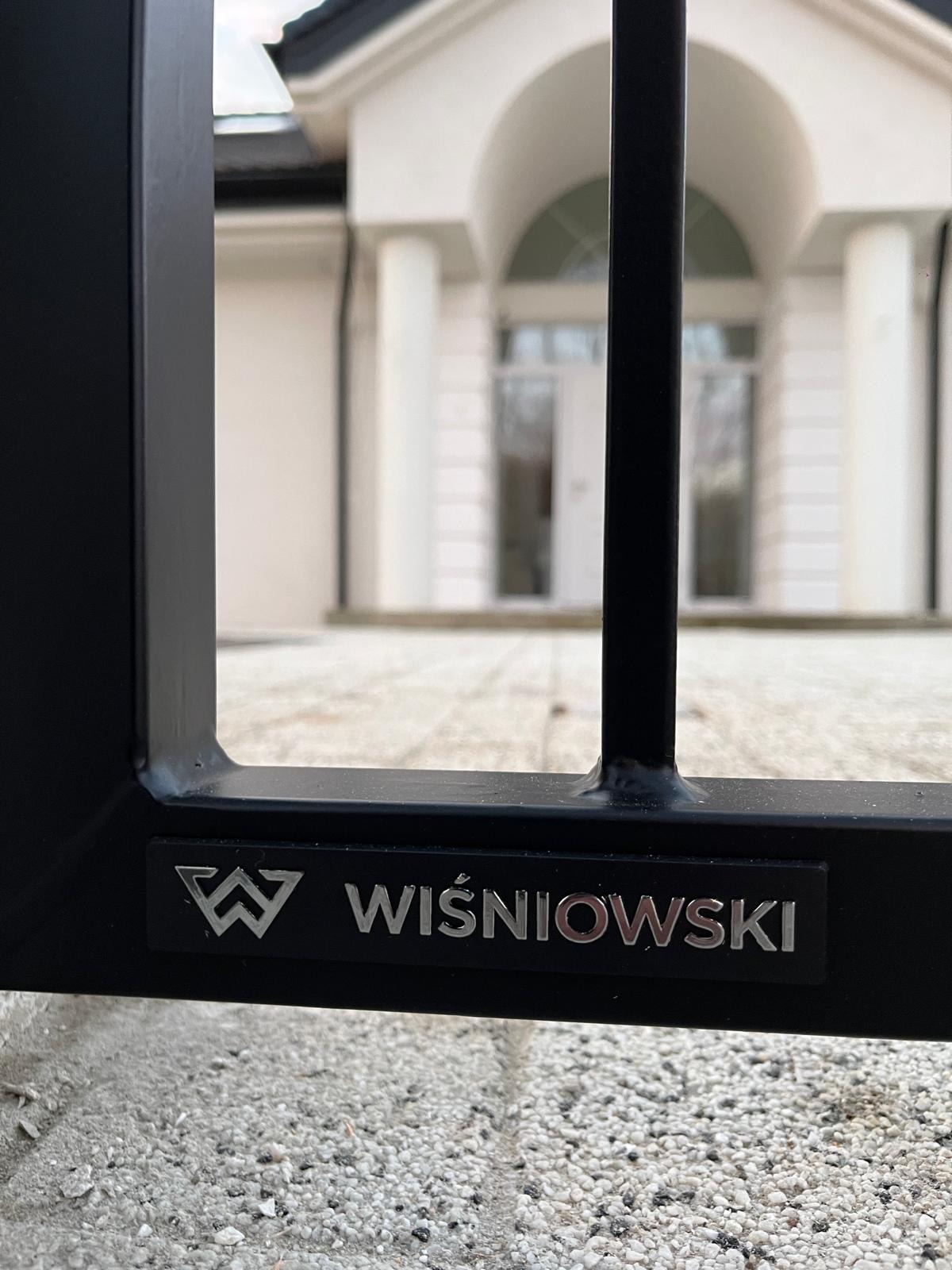 Fragment czarnej metalowej bramy Wiśniowski z widocznym logo na tle białego domu z kolumnami, ujęcie z dołu.