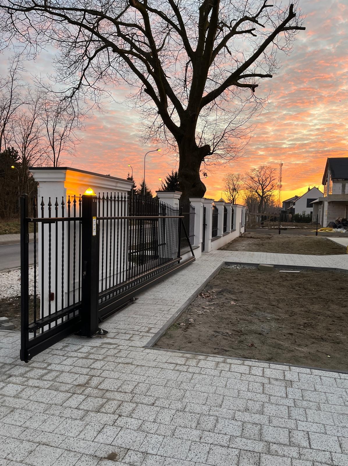 Otwarta, czarna brama wjazdowa z metalowymi prętami i białymi słupkami, prowadząca na posesję z kostką brukową. W tle drzewo bez liści i pomarańczowe niebo o zachodzie słońca.