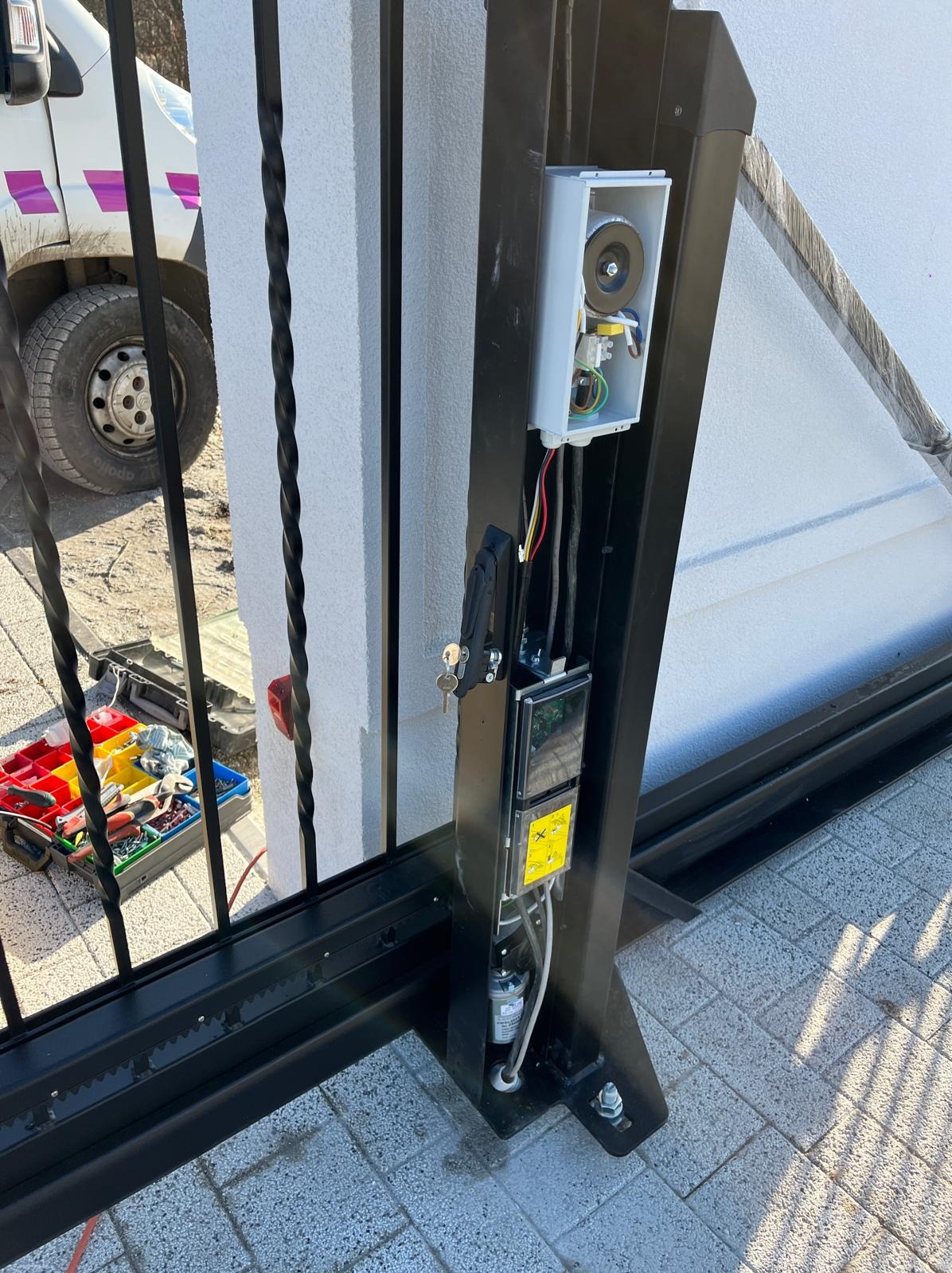 Czarna brama przesuwna z widoczną automatyką: otwartą obudową sterownika i elementami mechanicznymi. Obok skrzynka z narzędziami i fragment auta.