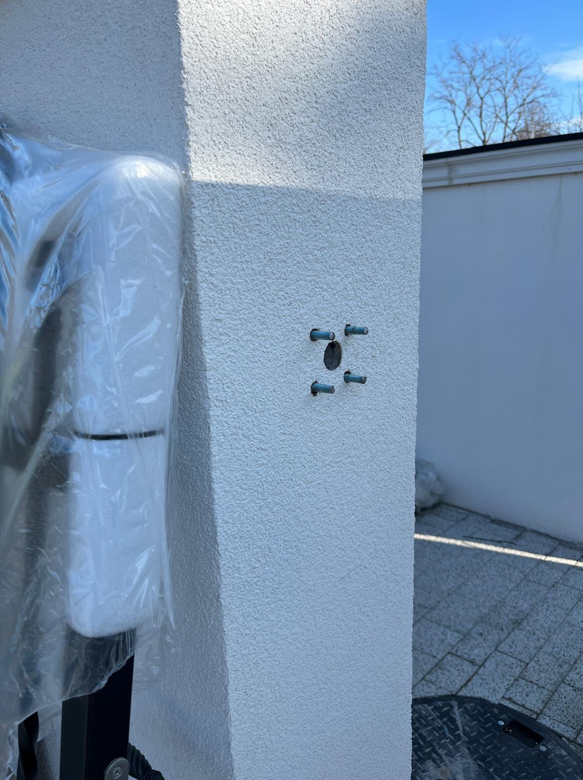 Przygotowanie do montażu automatu bramy: widoczne śruby w ścianie, obok automat w folii ochronnej, ściana o fakturze baranka, kostka brukowa.