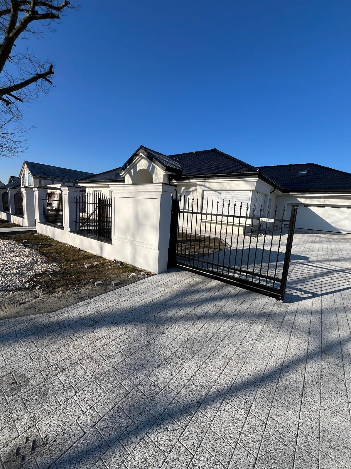 Nowoczesna, czarna brama wjazdowa z metalowymi prętami, osadzona w białych, murowanych słupkach. Widok na posesję z kostką brukową w słoneczny dzień.
