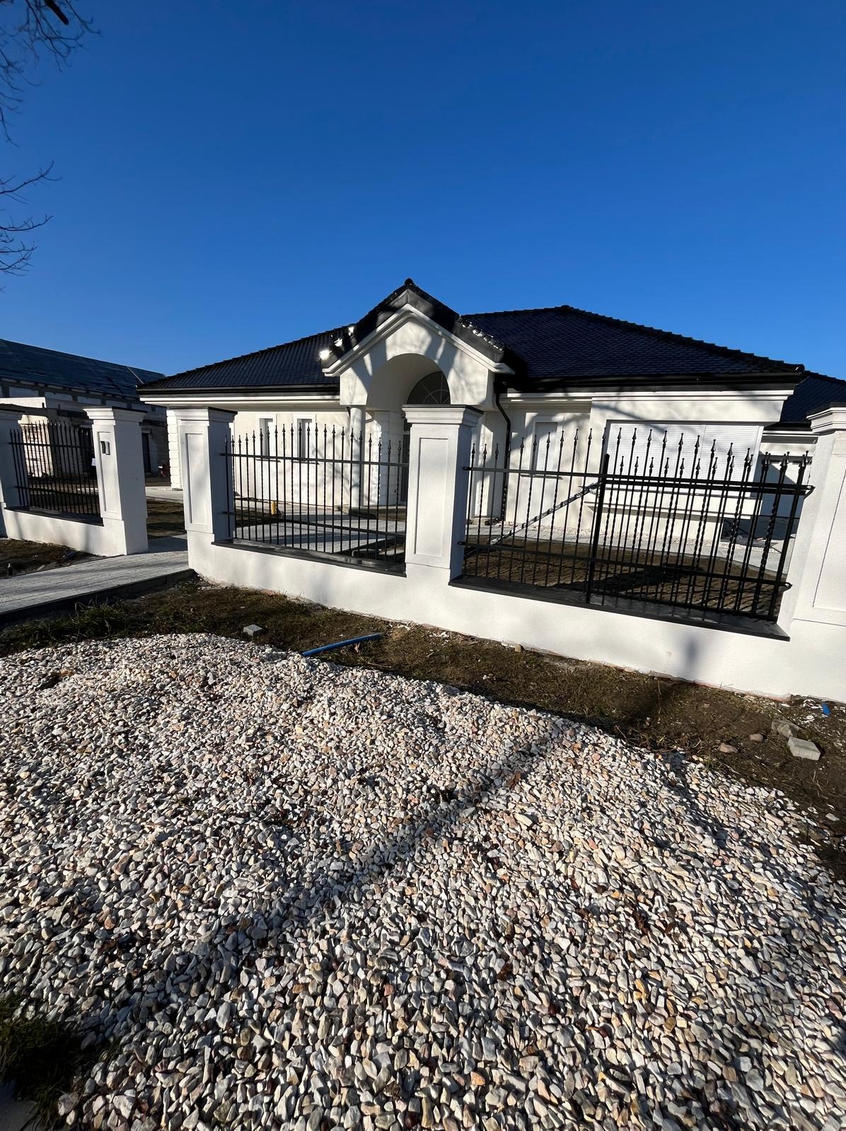 Eleganckie ogrodzenie metalowe z murowanymi słupkami, posesja w Mirkowie. Czarne, kute przęsła ogrodzeniowe na niskiej podmurówce. Dom w tle.