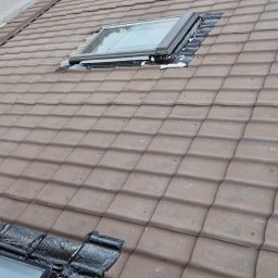 FULMAL Piotr Górka - Brązowa dachówka ceramiczna z oknem dachowym w trakcie uszczelniania czarną folią. Widoczne ślady po montażu i zabrudzenia na powierzchni dachu.